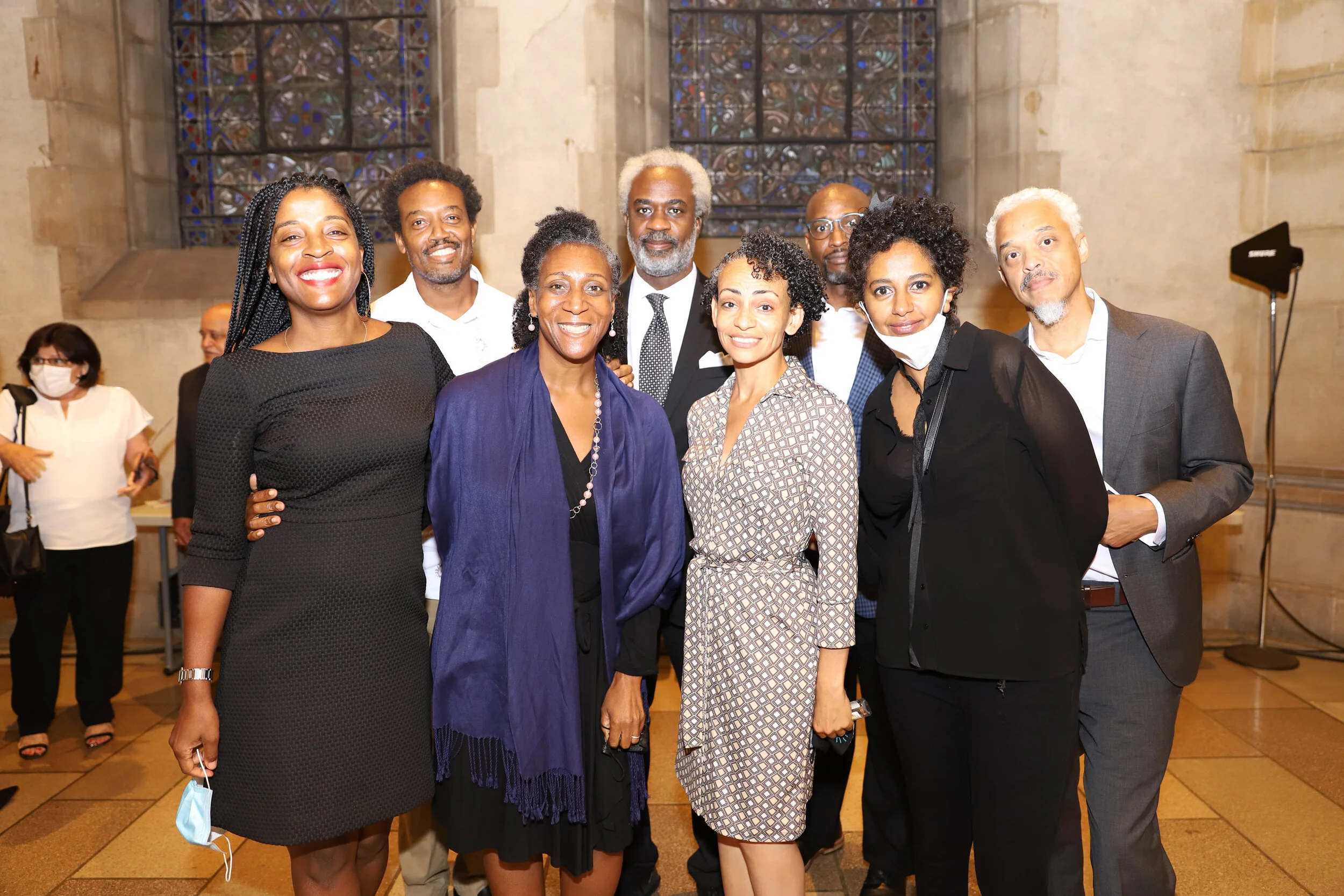Group of eight diverse adults smiling and standing together in a church with stained glass windows in the background.