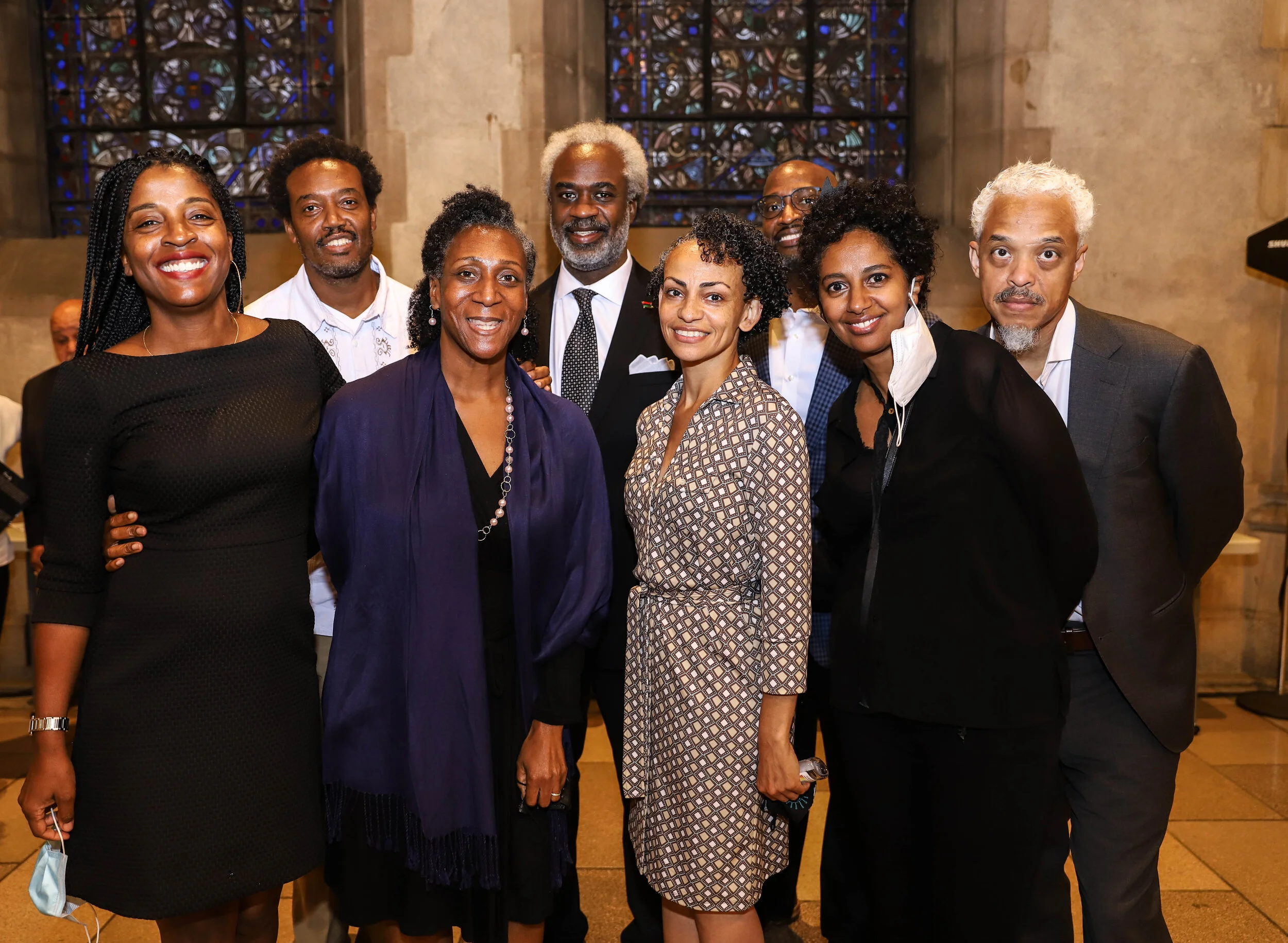 Group of nine diverse people smiling and posing together inside a building with stained glass windows in the background.