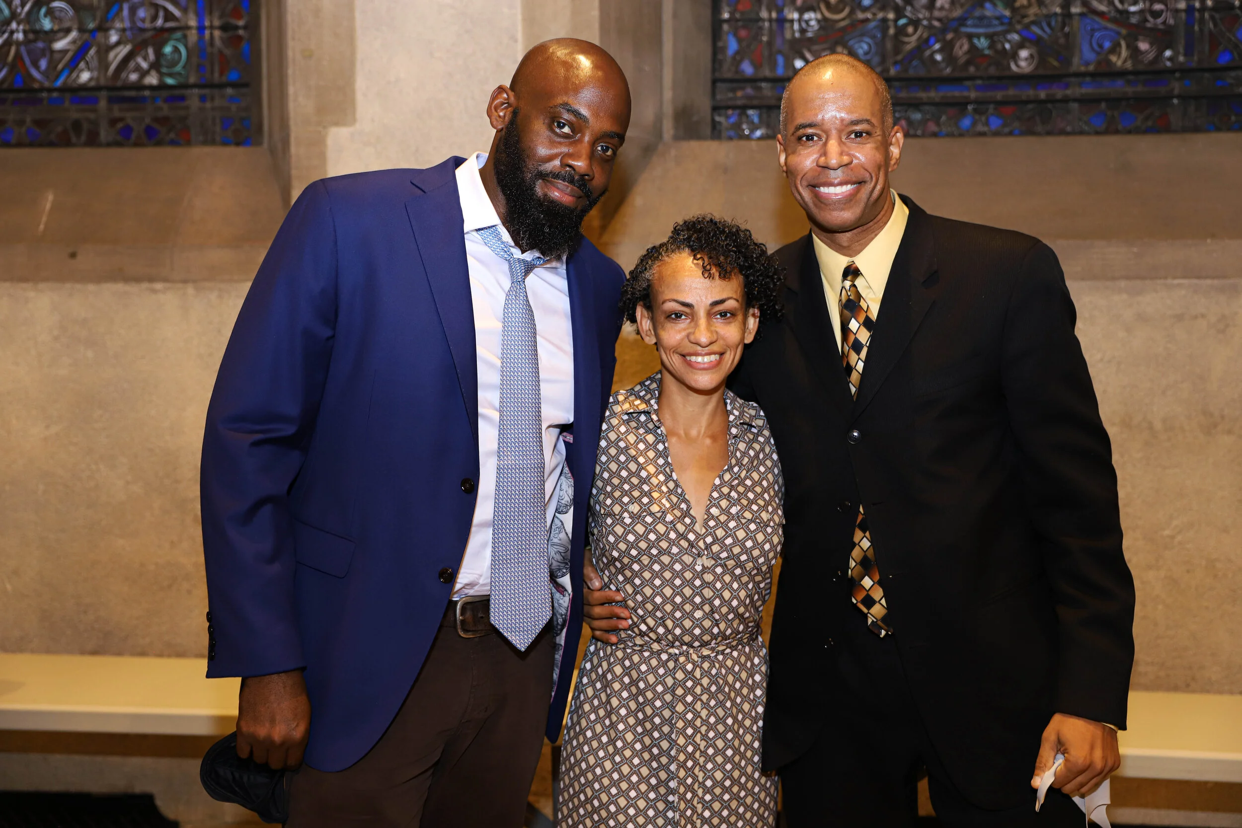 Three people standing indoors: a man in a blue suit with a white shirt and patterned tie, a woman in a patterned dress, and a man in a black suit with a yellow shirt and patterned tie. They are smiling and posing for the photo, with colorful stained 