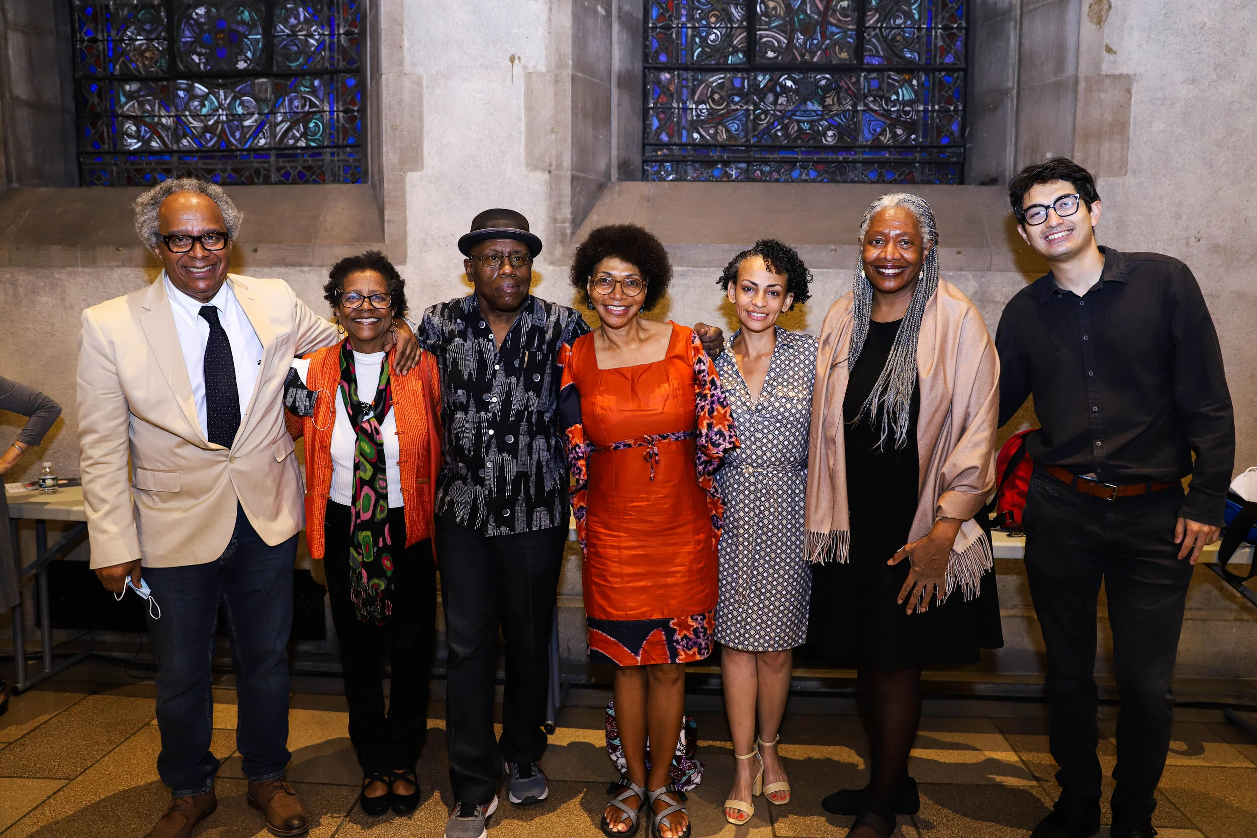 A diverse group of eight people standing together indoors in front of stained glass windows, smiling at the camera.
