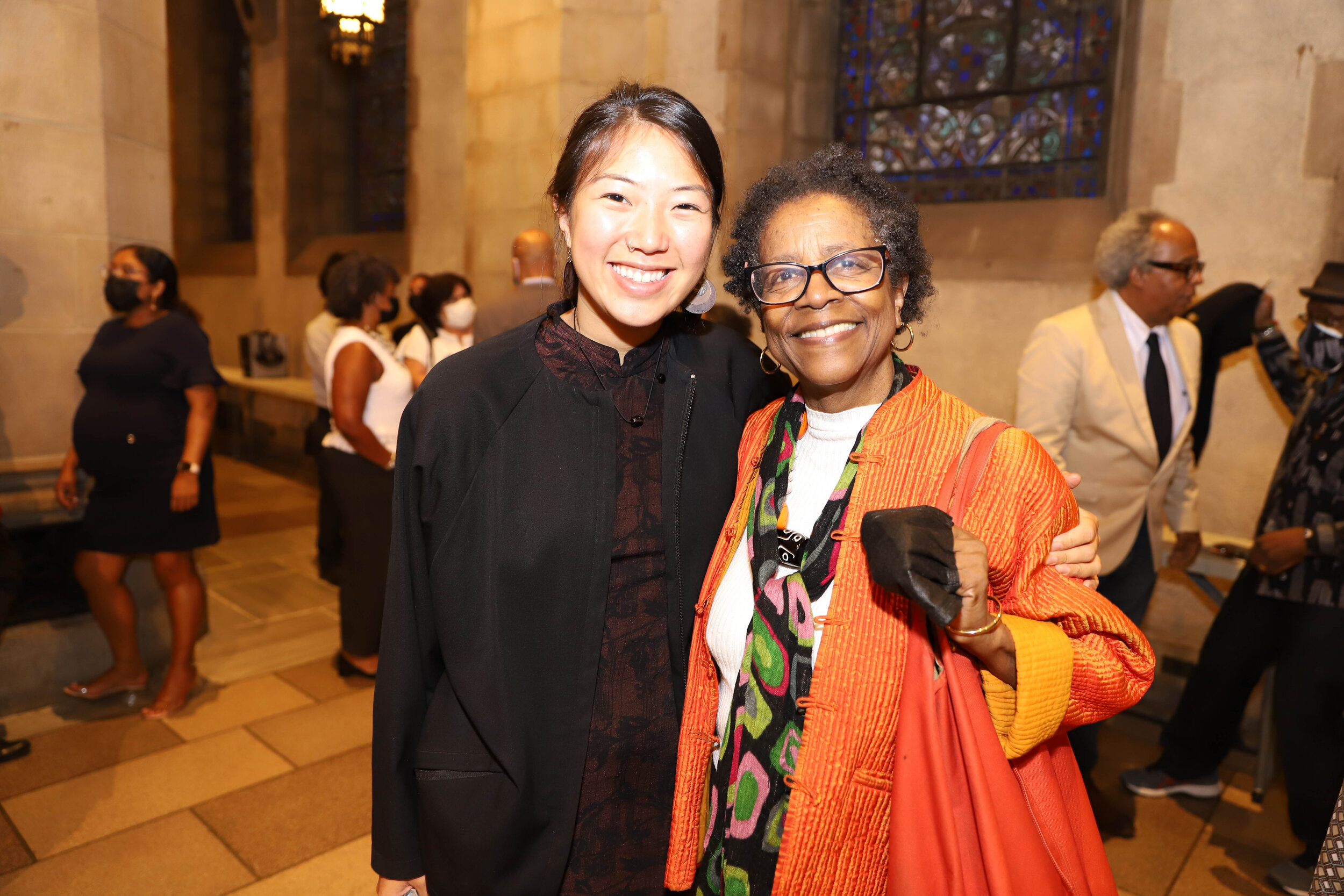 Two women smiling and hugging each other inside a large church or cathedral with stained glass windows. One woman has light skin and dark hair, and the other has dark skin and gray hair.