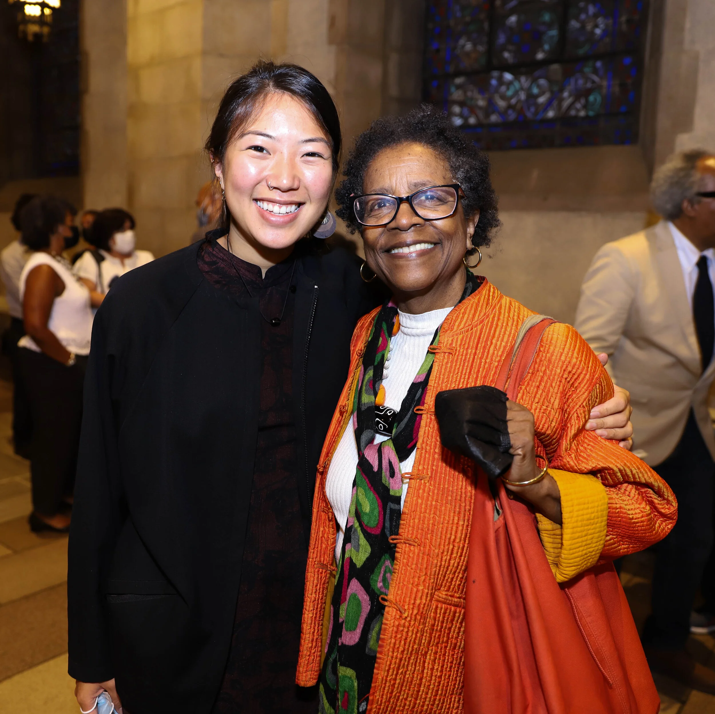 Two women smiling and embracing inside a church with stained glass windows in the background.