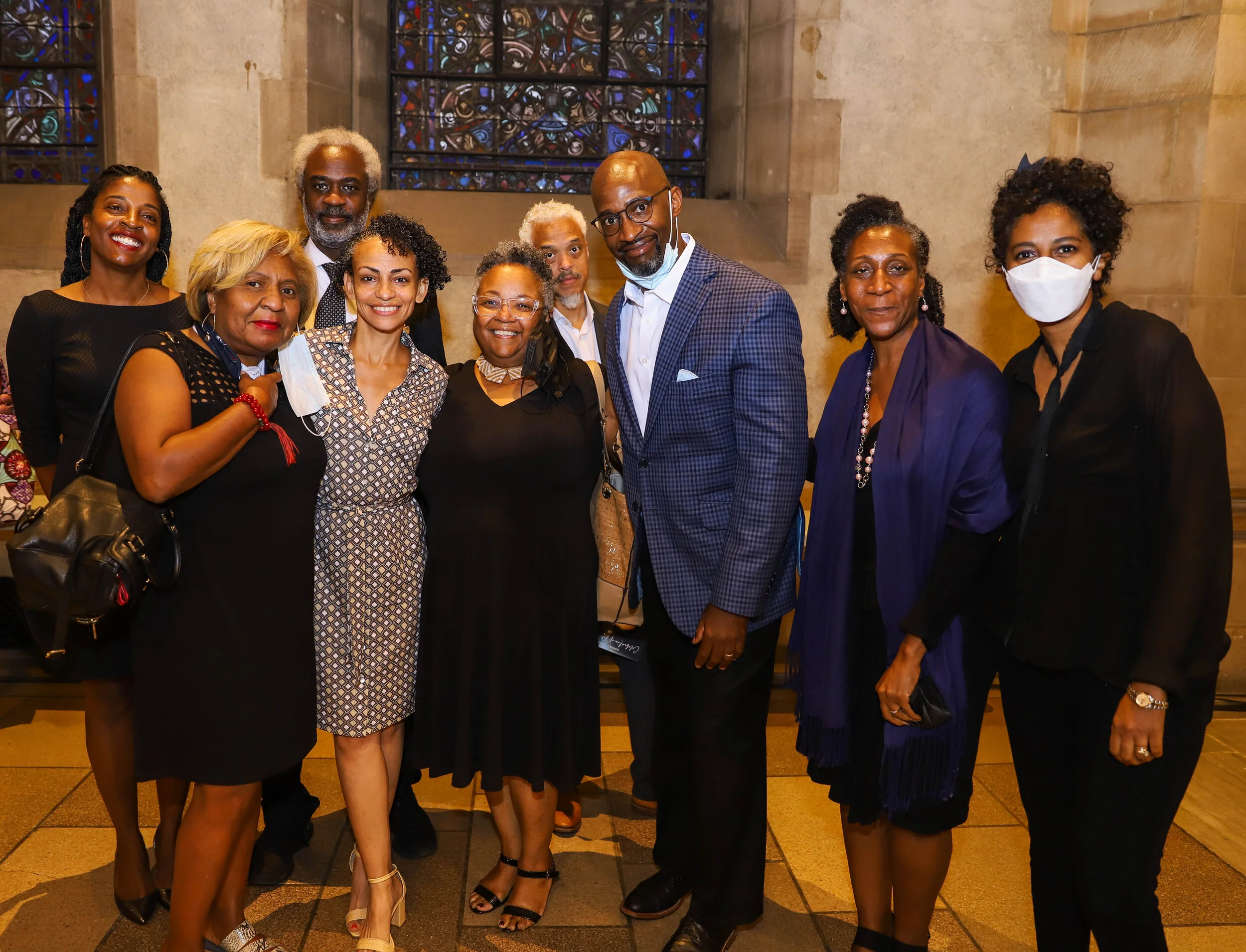 A diverse group of ten people, including men and women of different ages, poses together inside a church or cathedral with stained glass windows in the background. Some are smiling, one woman is wearing a face mask, and they are dressed in semi-forma