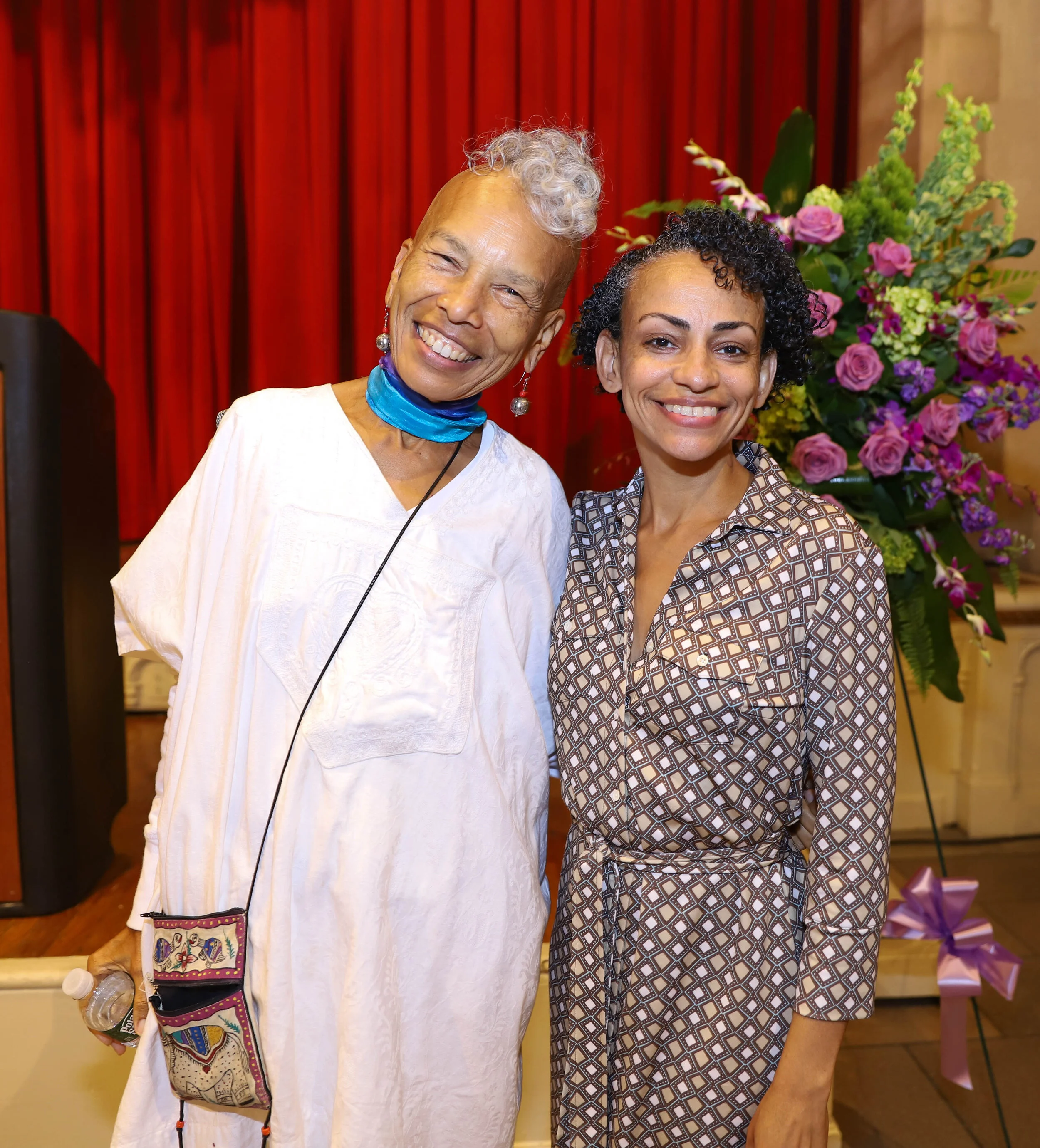 Two women smiling in front of a red curtain with a large flower arrangement in the background. One woman is older, with short curly gray hair, wearing a white top, and a blue face mask hanging below her chin. The other woman is younger, with short bl