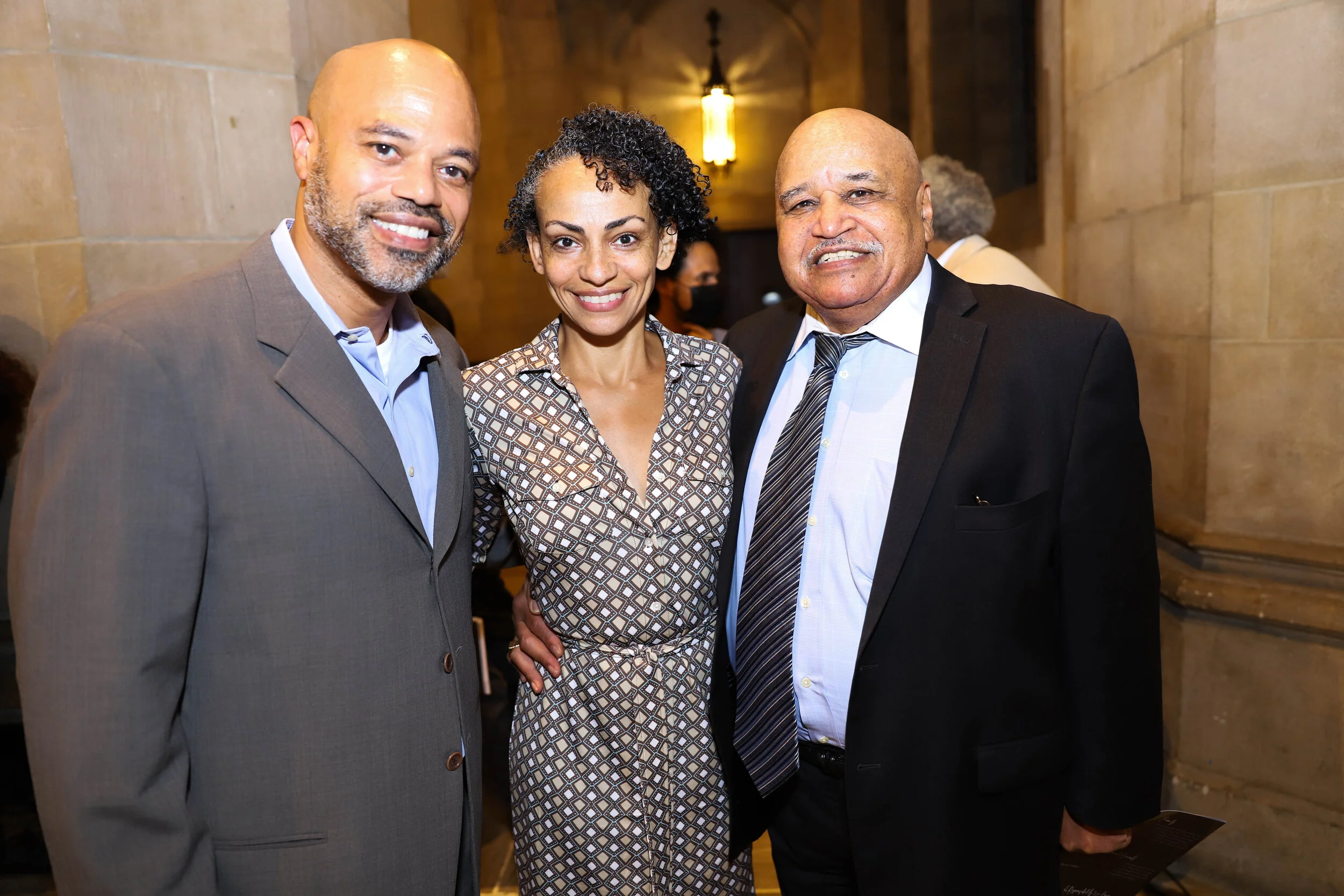 Three people standing together at an event, smiling at the camera. Two men are in suits, and a woman is in a patterned dress. The setting is indoors with stone walls and warm lighting.