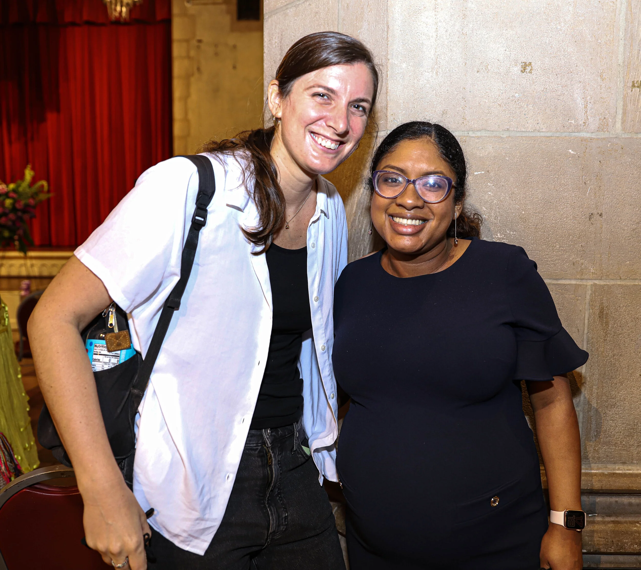 Two women standing close together, smiling, in front of a stone wall inside a building. One woman has long brown hair, light skin, and is wearing a white shirt. The other woman has dark hair, darker skin, and purple glasses, and is wearing a black to