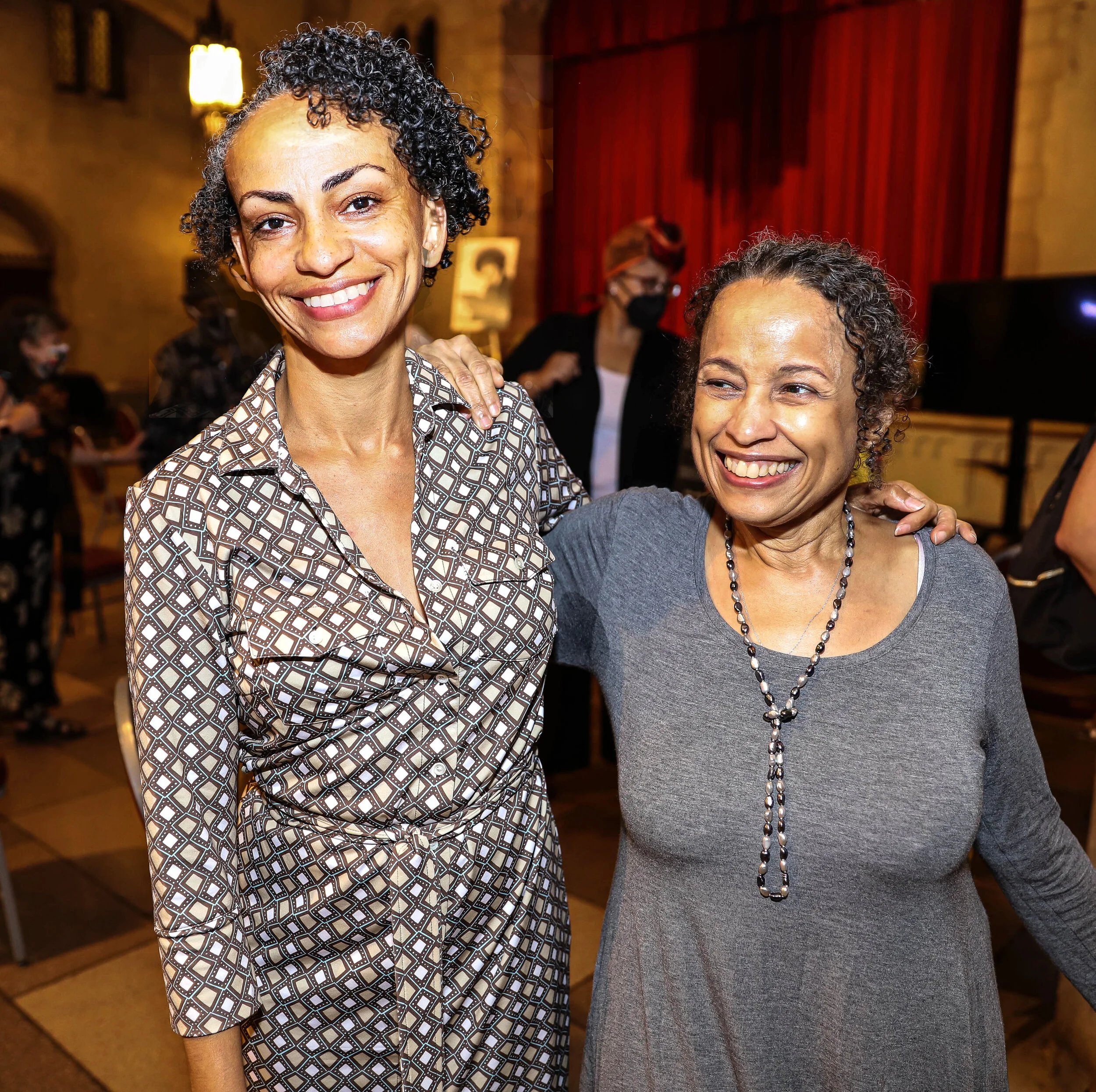 Two women smiling and posing together at an event, with red curtains and warm lighting in the background.