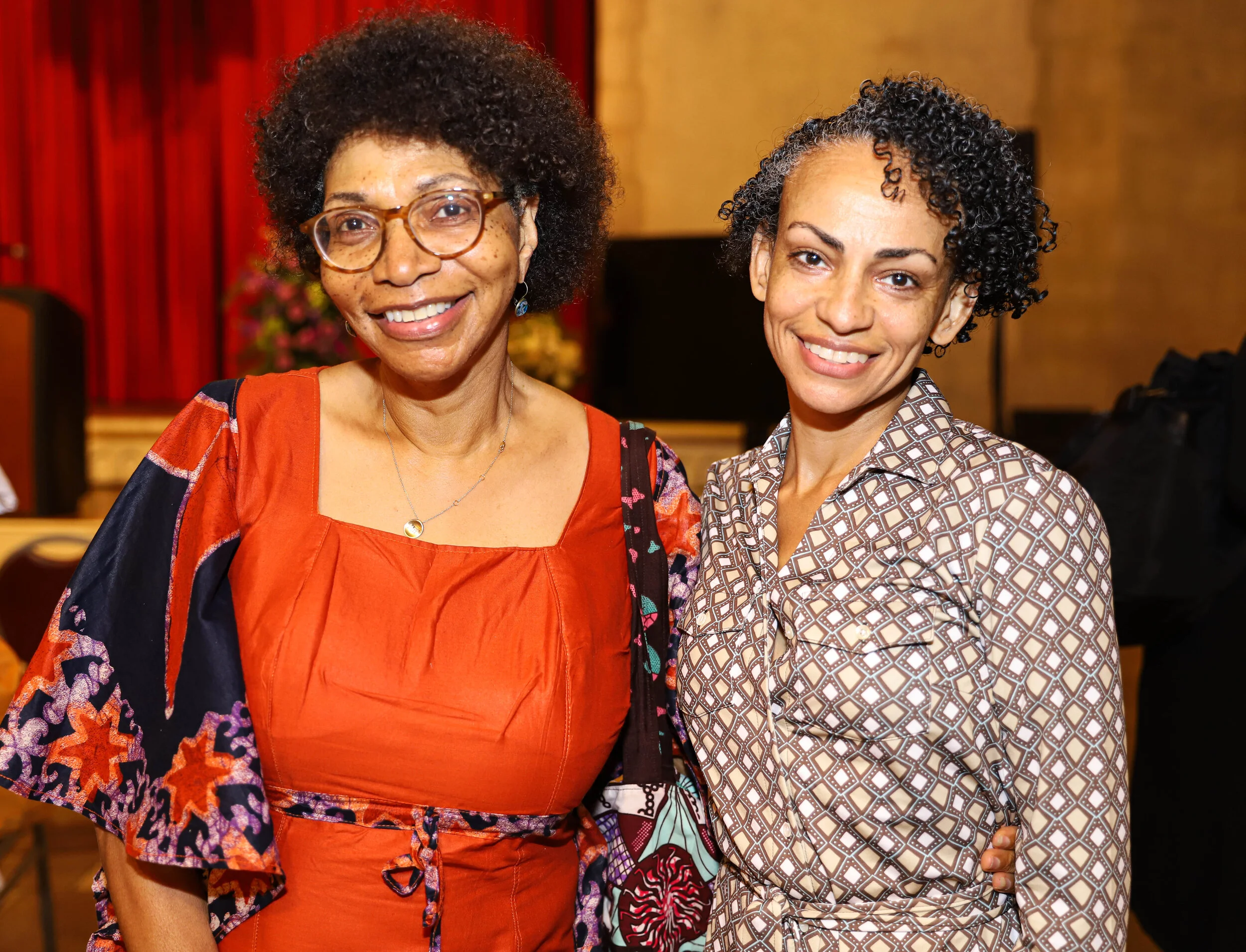 Two smiling women, one with glasses and short curly hair wearing a patterned jacket over a beige shirt, and the other with short curly black hair wearing a red and black patterned dress, standing indoors with a red curtain and beige wall background.