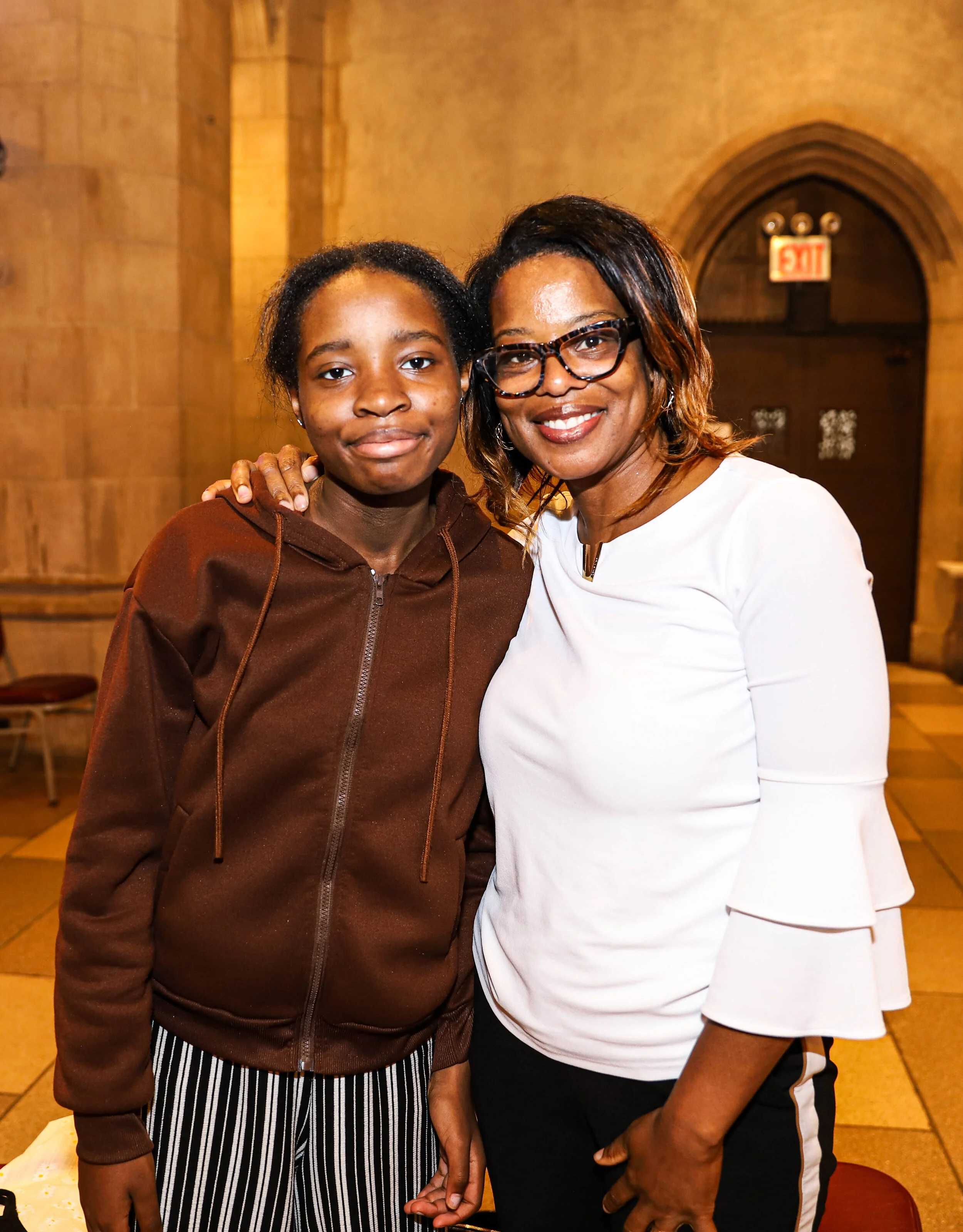 A woman and a girl stand together indoors in front of a wooden wall, smiling and holding hands, with the woman wearing glasses and a white top, and the girl wearing a brown hoodie and striped pants.