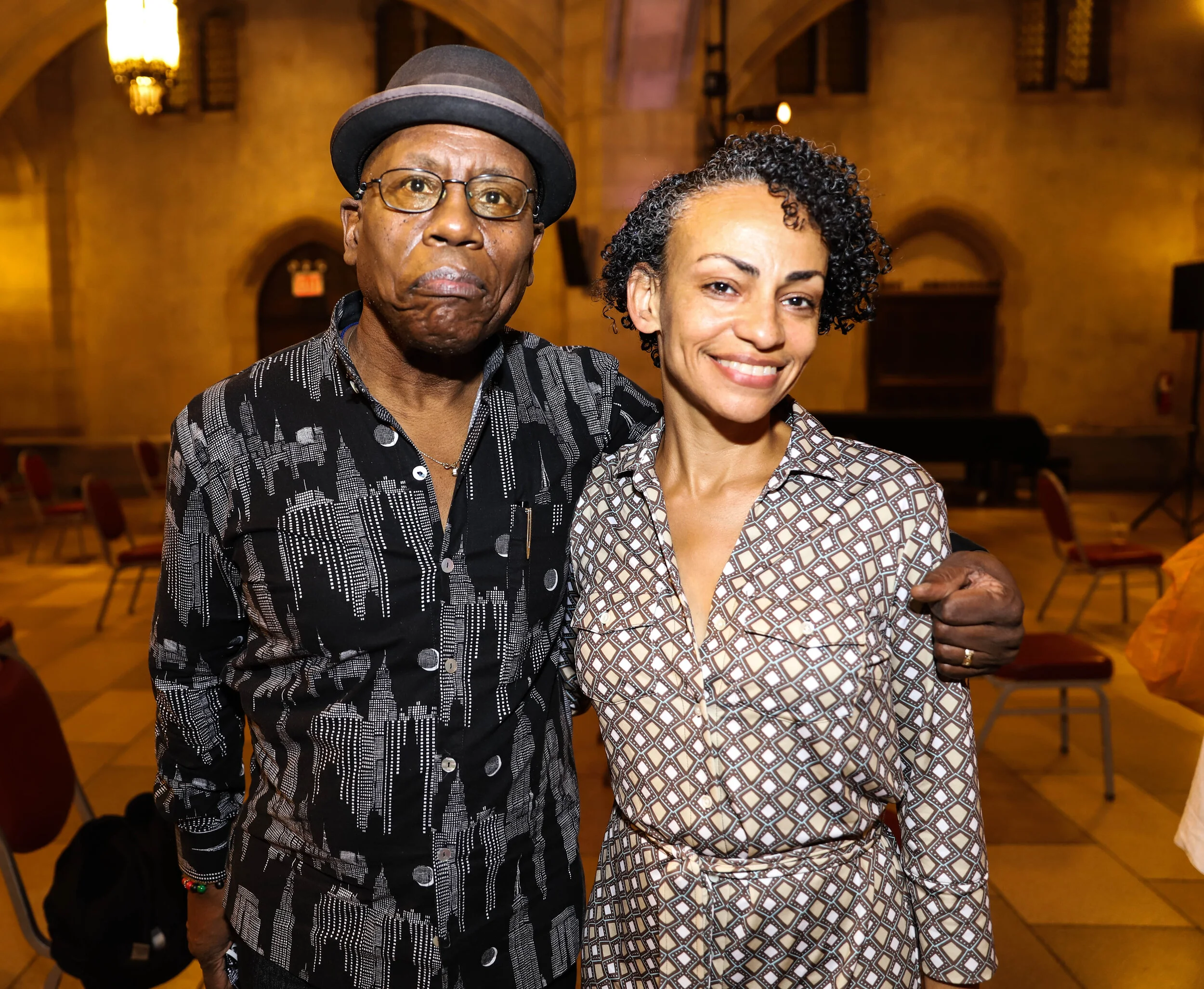 A man and woman standing together in an interior setting with brick walls, arched doorways, and chairs in the background. The man wears glasses, a hat, and a printed shirt, while the woman has short curly hair and is smiling, wearing a patterned shir