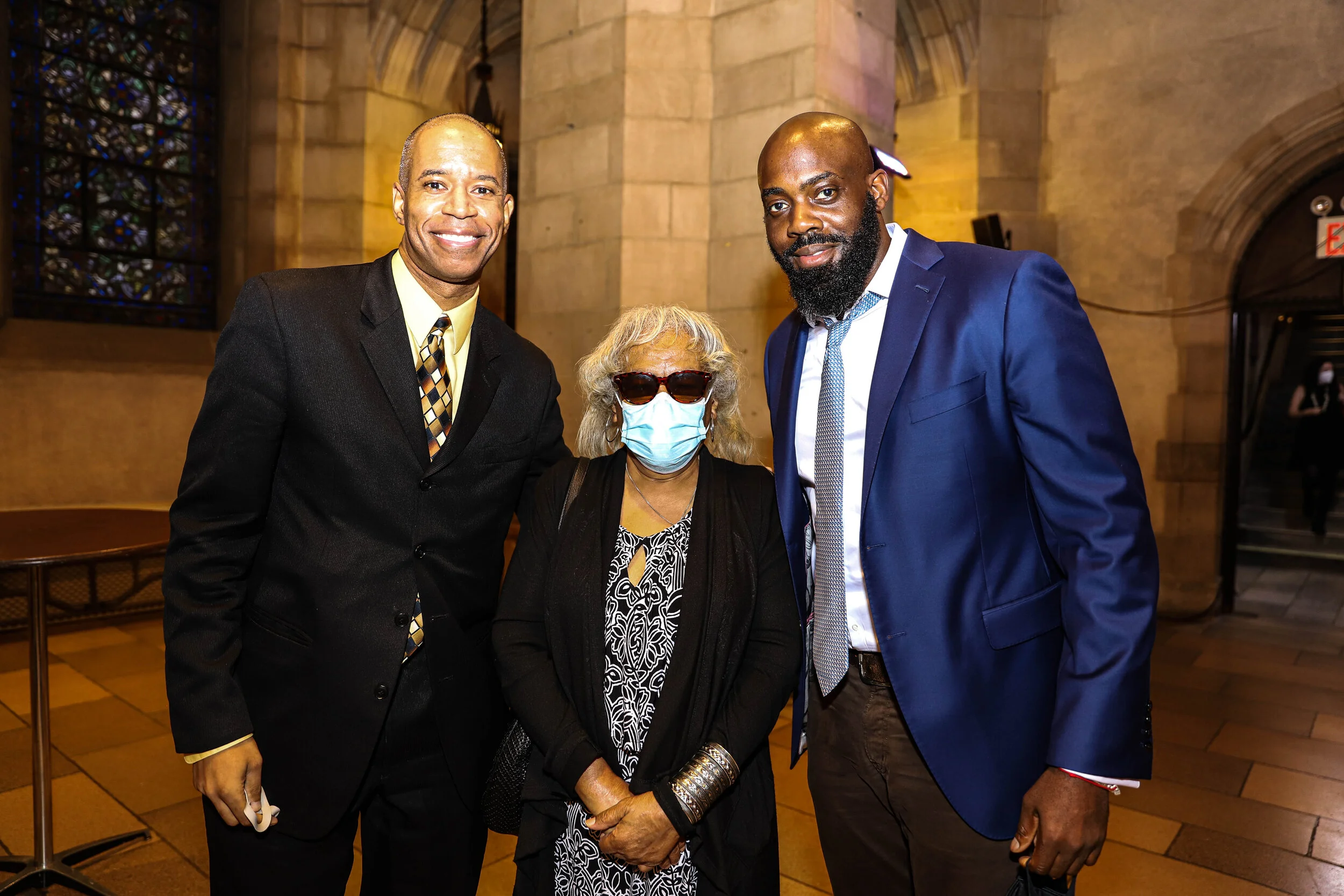 Four people standing inside a church. Two men are wearing suits, a woman is wearing sunglasses and a face mask, and another man is partially visible on the right, also in formal attire.