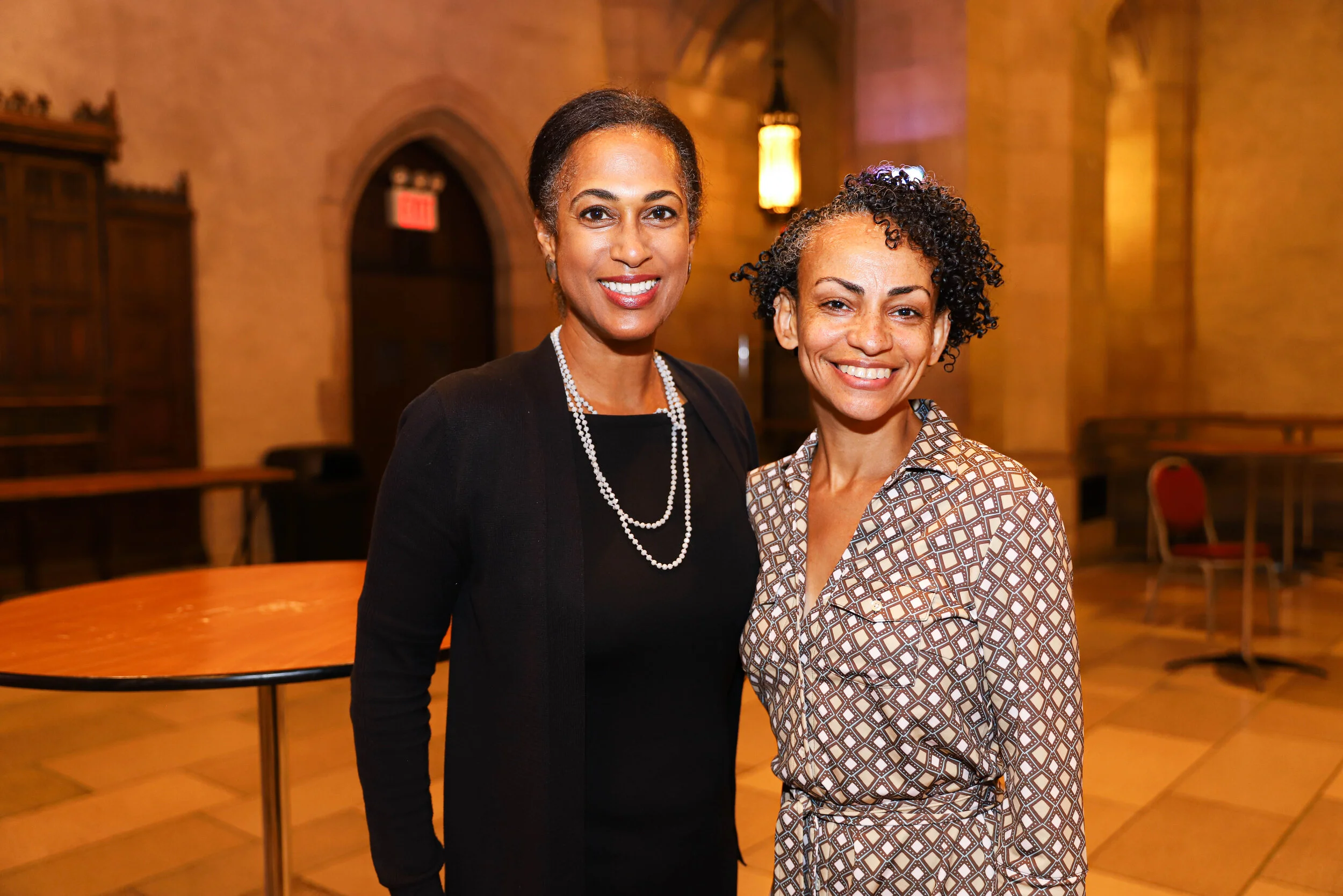 Two women smiling indoors, one wearing a black top with a pearl necklace and the other in a patterned shirt. Warm lighting and wooden decor are in the background.