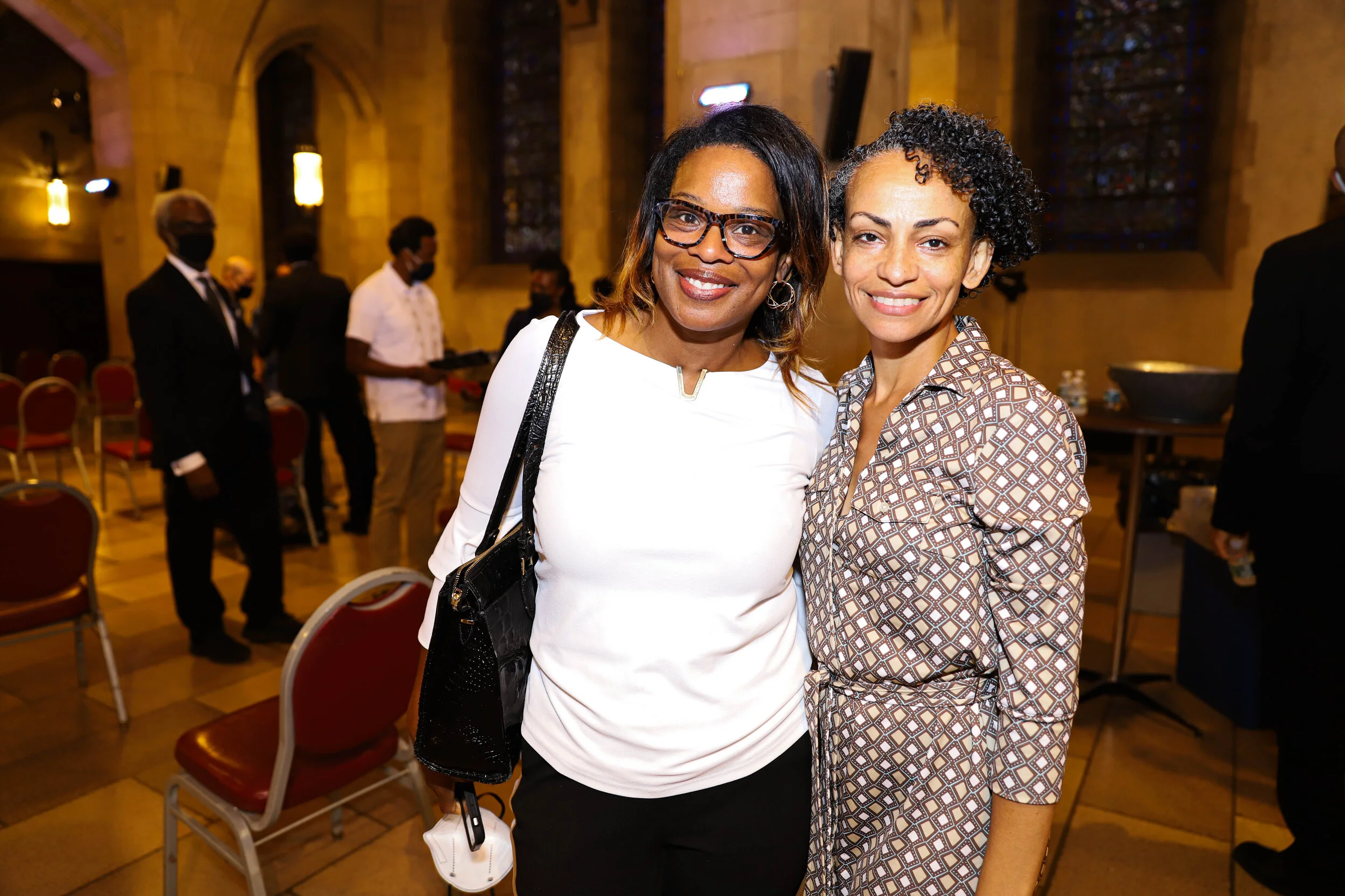 Two women smiling and posing for a photo inside a church or hall with stained glass windows in the background. They are dressed in semi-formal attire, and other people are visible in the background.