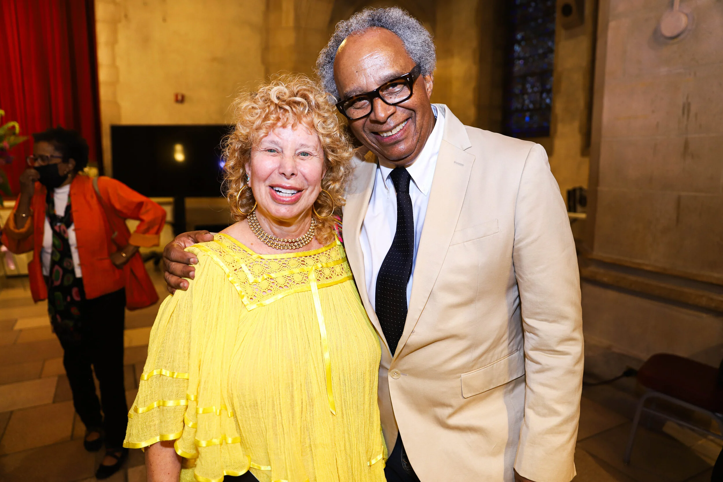 Two people smiling and posing for a photo at an indoor event. The woman on the left is wearing a bright yellow dress with lace details and jewelry, and the man on the right is dressed in a light beige suit with a white shirt and black tie. There is a