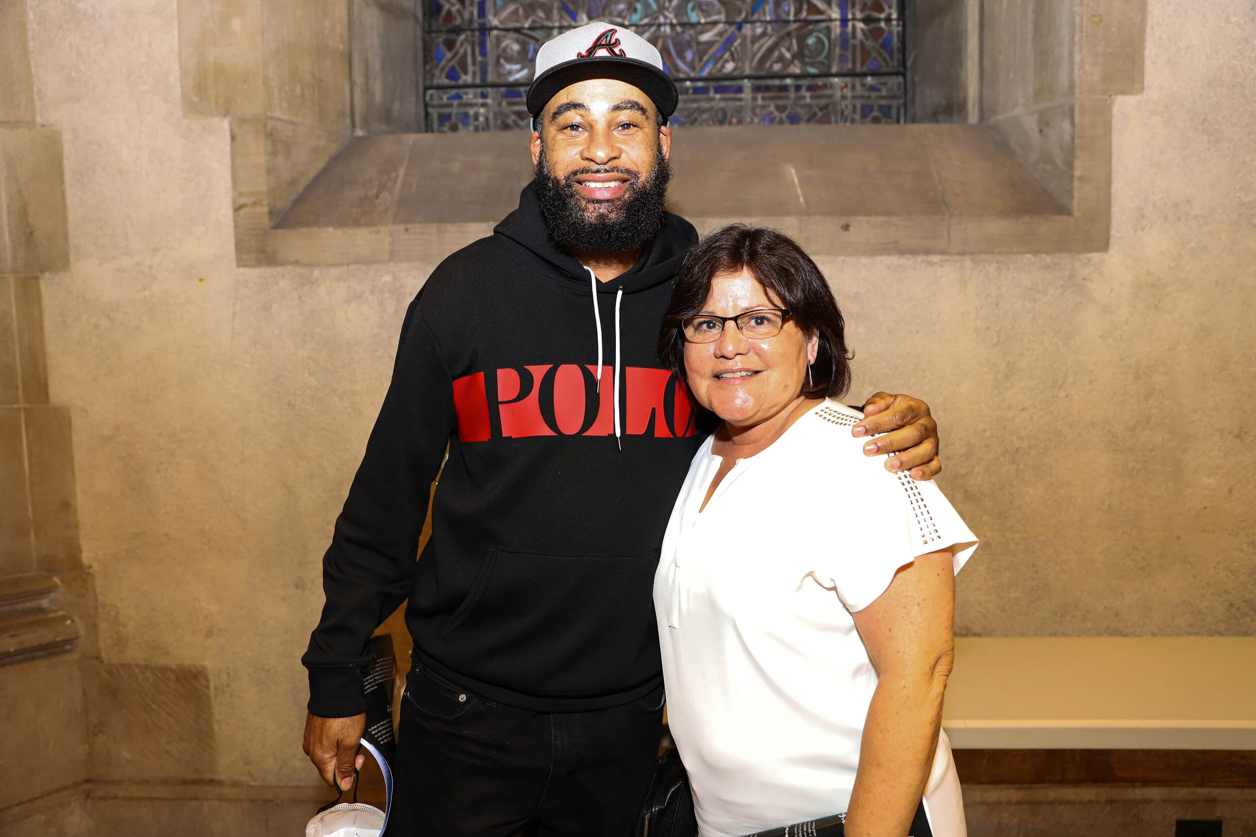 A tall man with a beard wearing a grey baseball cap with a red and black logo, a black hoodie with red lettering, and black pants standing indoors next to a woman with short dark hair, glasses, and a white blouse. They are smiling and have their arms