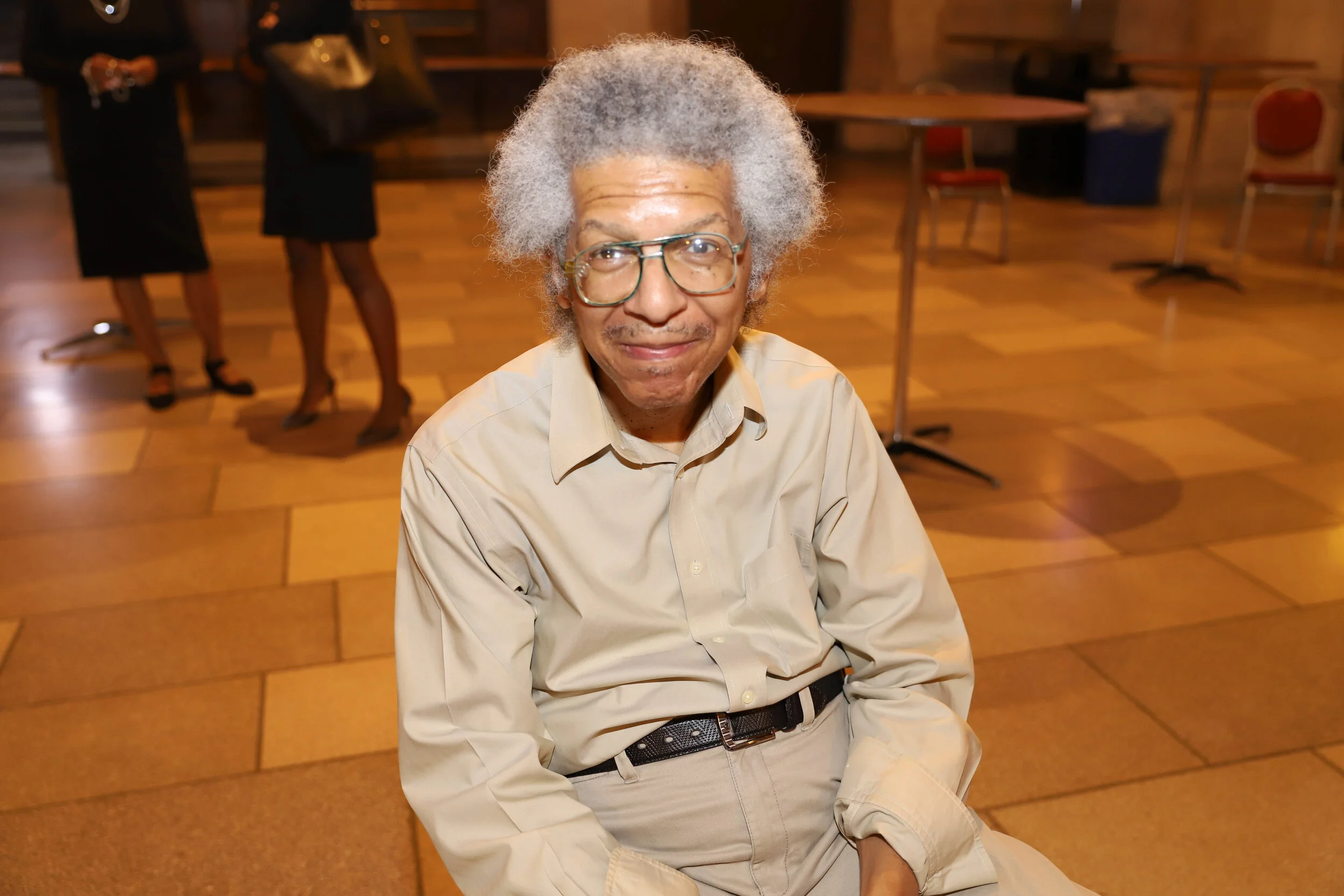 An elderly man with gray hair and glasses sitting on the floor in a room with wooden floors and tables, smiling at the camera. In the background, two women are standing and talking, one holding a cup.