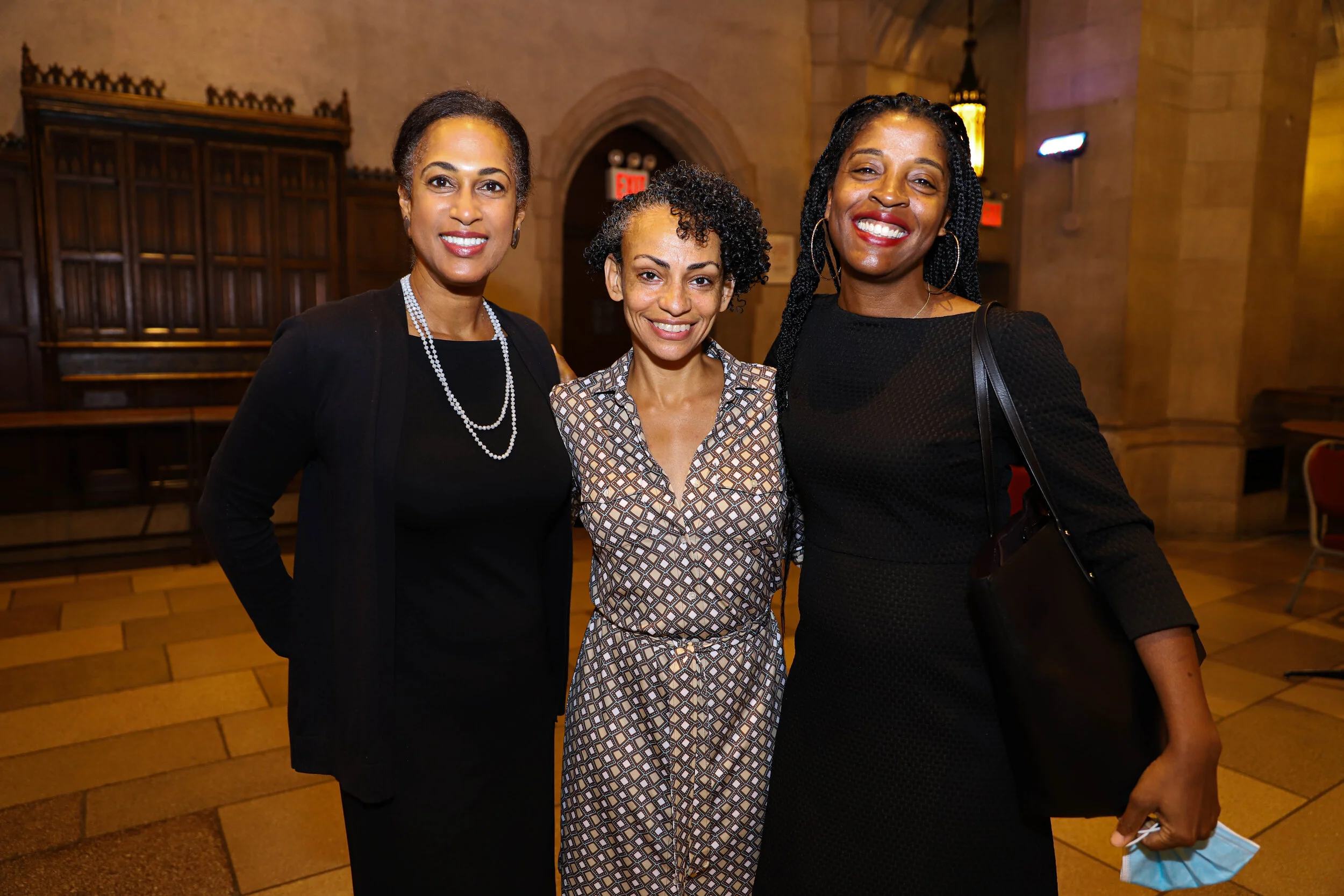 Three women standing close together and smiling at the camera inside a historic-style building with stone walls and hardwood floors.