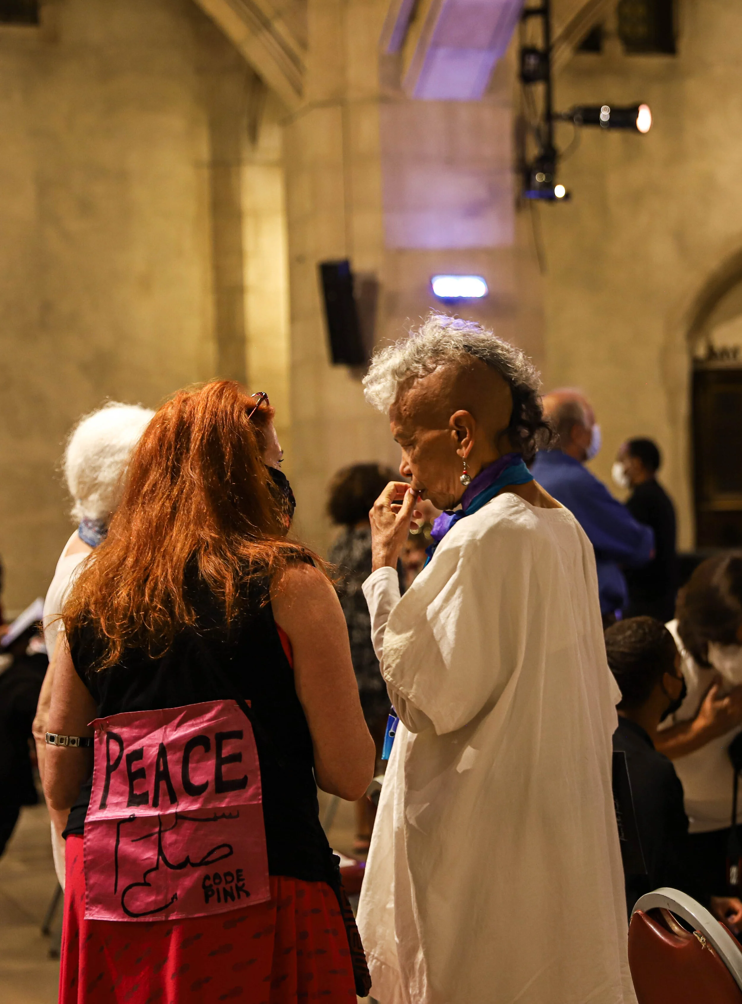 Two women are engaged in a conversation in a crowd indoors. One woman has long red hair and is wearing a black top with a pink bag that has the word PEACE on it. The other woman has short curly gray hair, earrings, a purple scarf, and a cream-colored