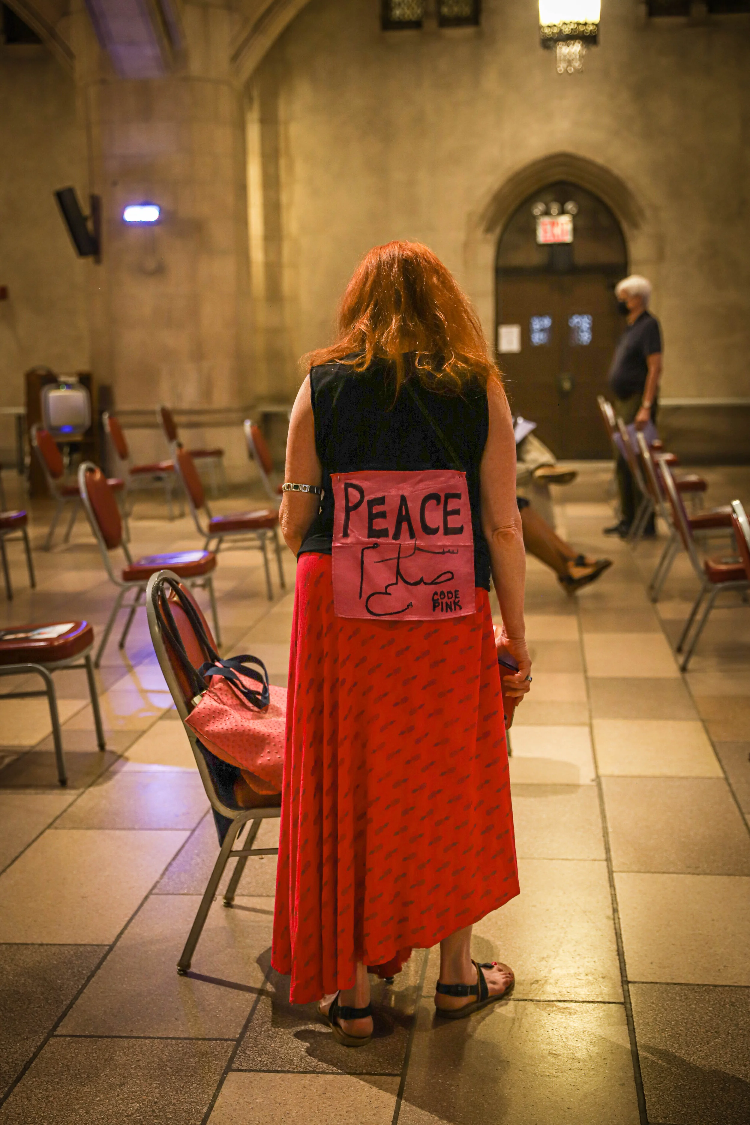 A woman with red hair wearing a black top with a peace sign and a red skirt with small black patterns, standing in a room with chairs and a person in the background. The room has stone walls, arched doorways, and stained glass windows.