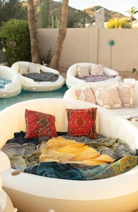 Outdoor pool area with bubble-shaped lounge chairs and colorful pillows, some covered with blankets, surrounded by trees and a beige wall.