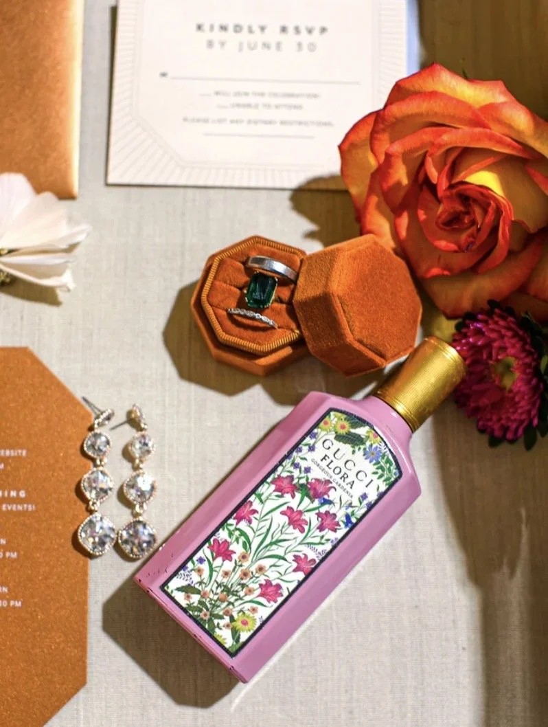 A flat lay of a gift or invitation with a Gucci Flora perfume bottle, jewelry including earrings and a ring in a velvet box, a large rose, and other flowers on a neutral fabric surface.