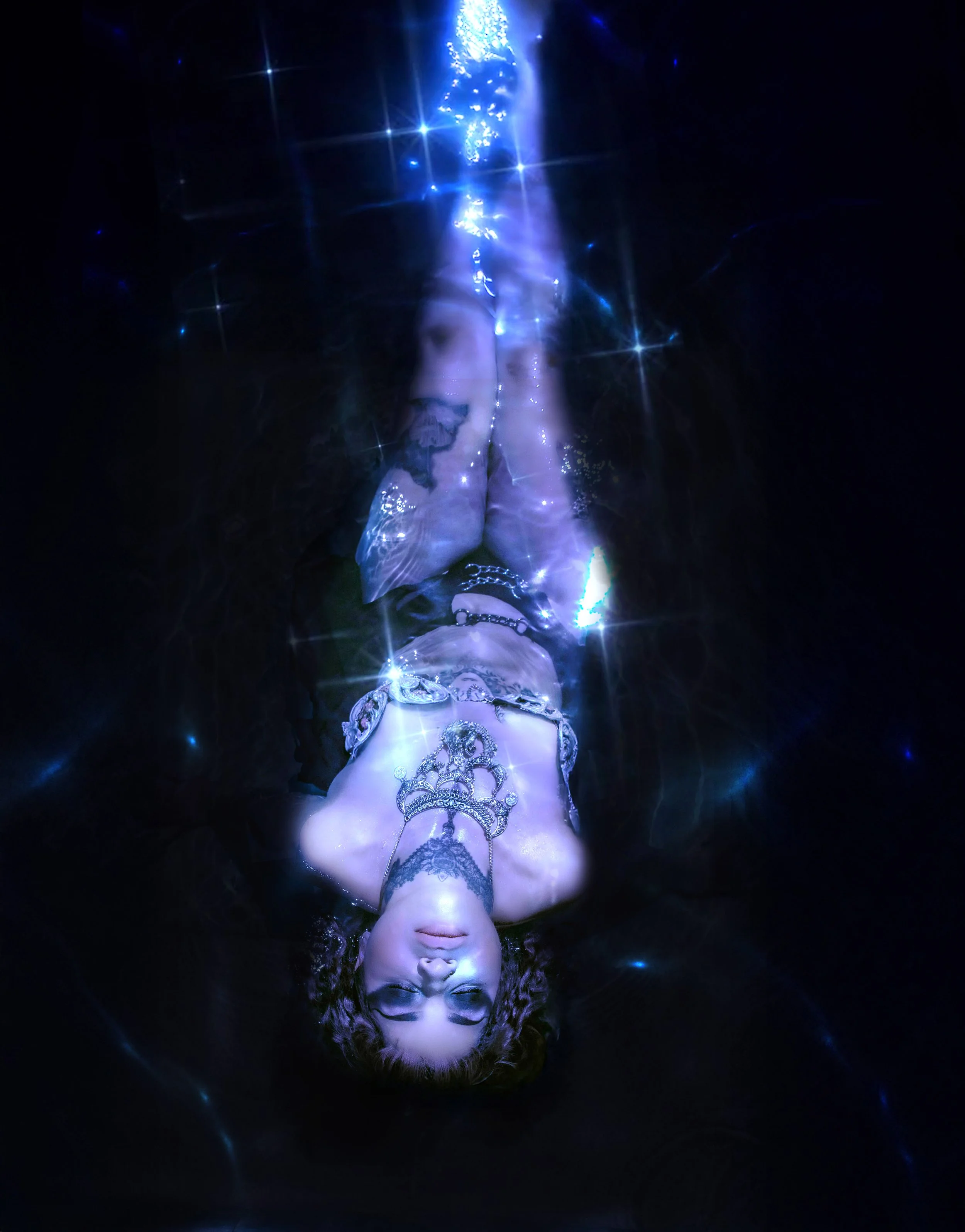 A woman lying on her back with her eyes closed, illuminated by blue and purple lights, surrounded by star-like reflections, wearing a decorative jewelry piece on her forehead and chest, against a dark background.
