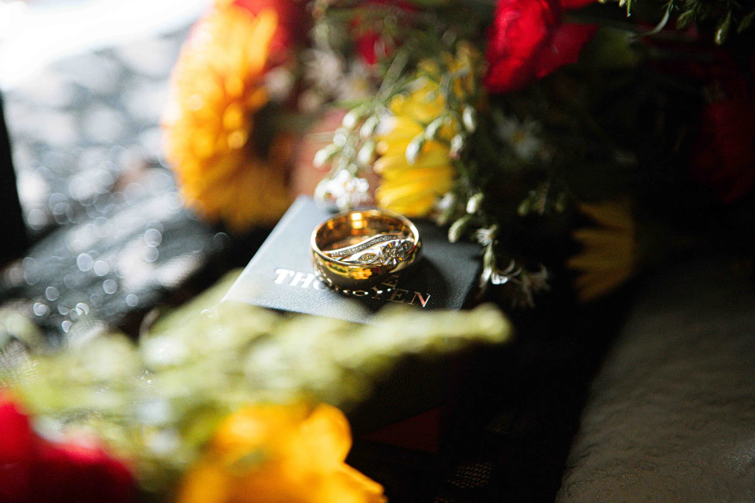 Close-up photograph of wedding rings displayed among colorful flowers and warm candlelight.
