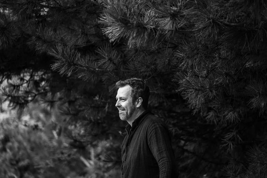 A man with a beard and short hair smiling and looking to the left, standing outdoors in front of a large pine tree.
