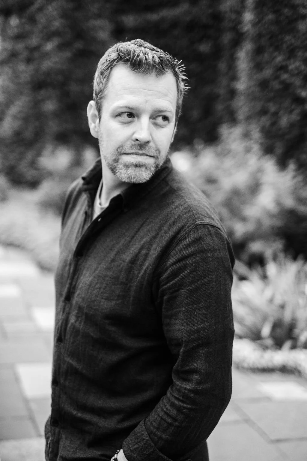 Black and white portrait of a man with short wavy hair and a beard, wearing a dark long-sleeved shirt, standing outdoors with blurred trees and plants in the background, looking to his left.
