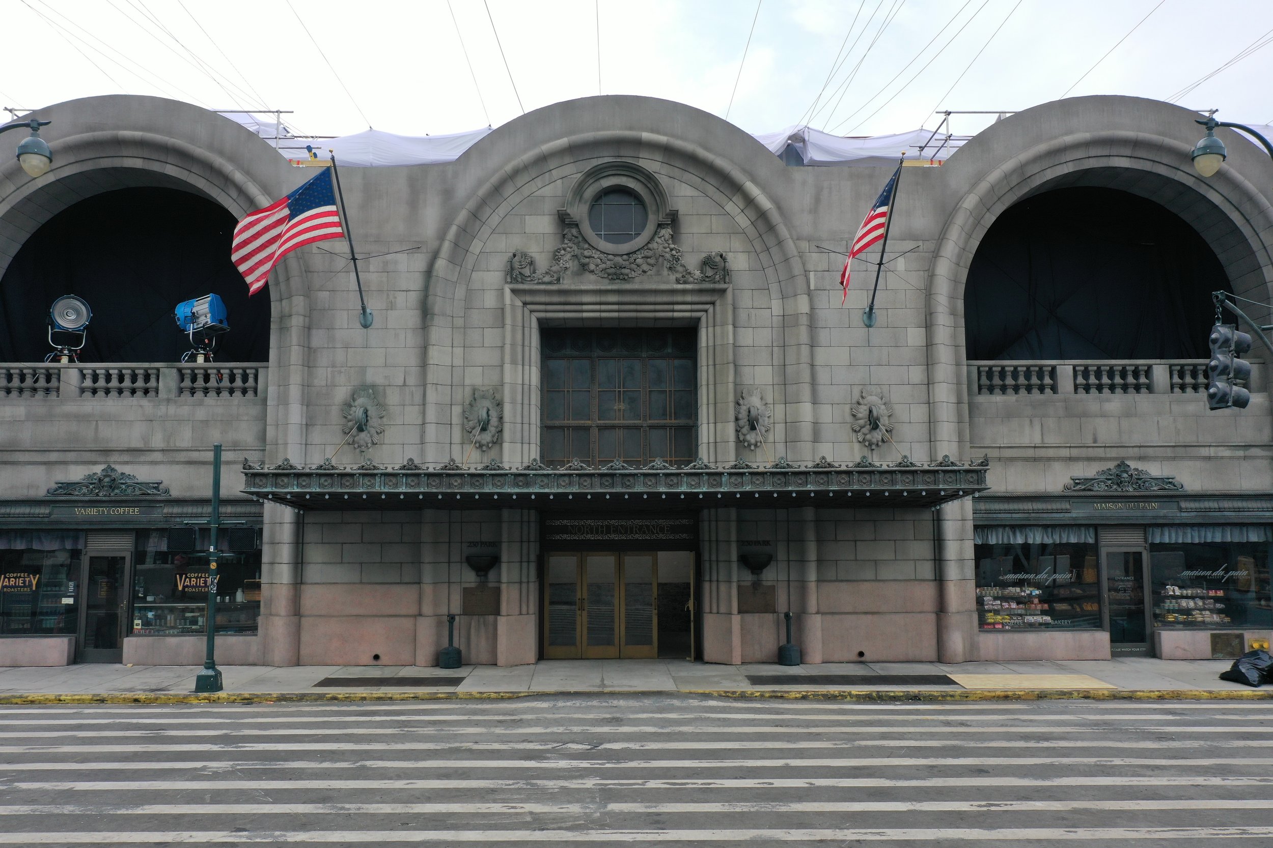 Front view of a historic building with two American flags, decorative stonework, and a marquee that reads 'North Entrance.' The building has large windows and is flanked by storefronts, with a crosswalk and street in the foreground.