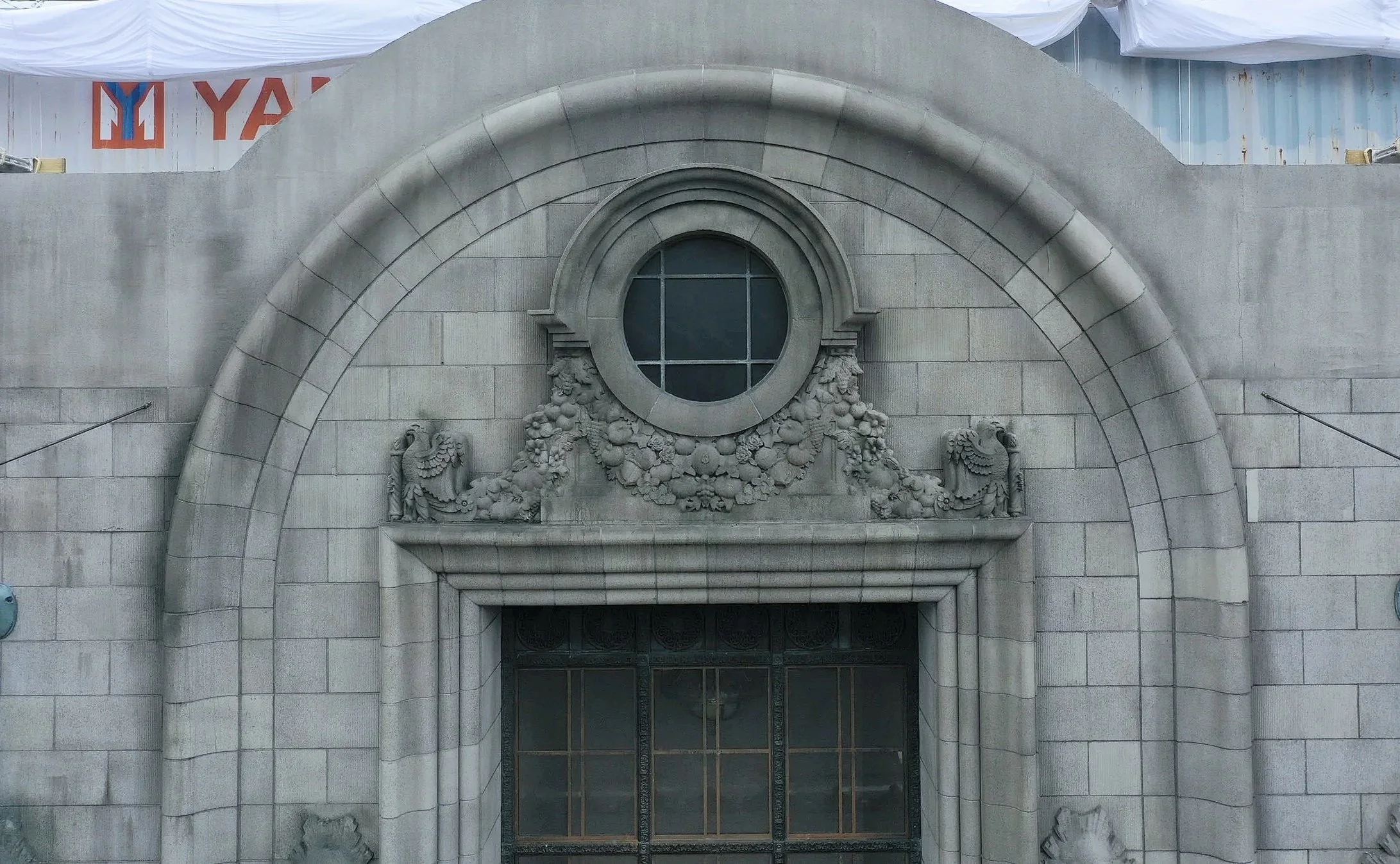 Close-up of an ornate stone building facade with a circular window and decorative floral and eagle reliefs above a metal gate.