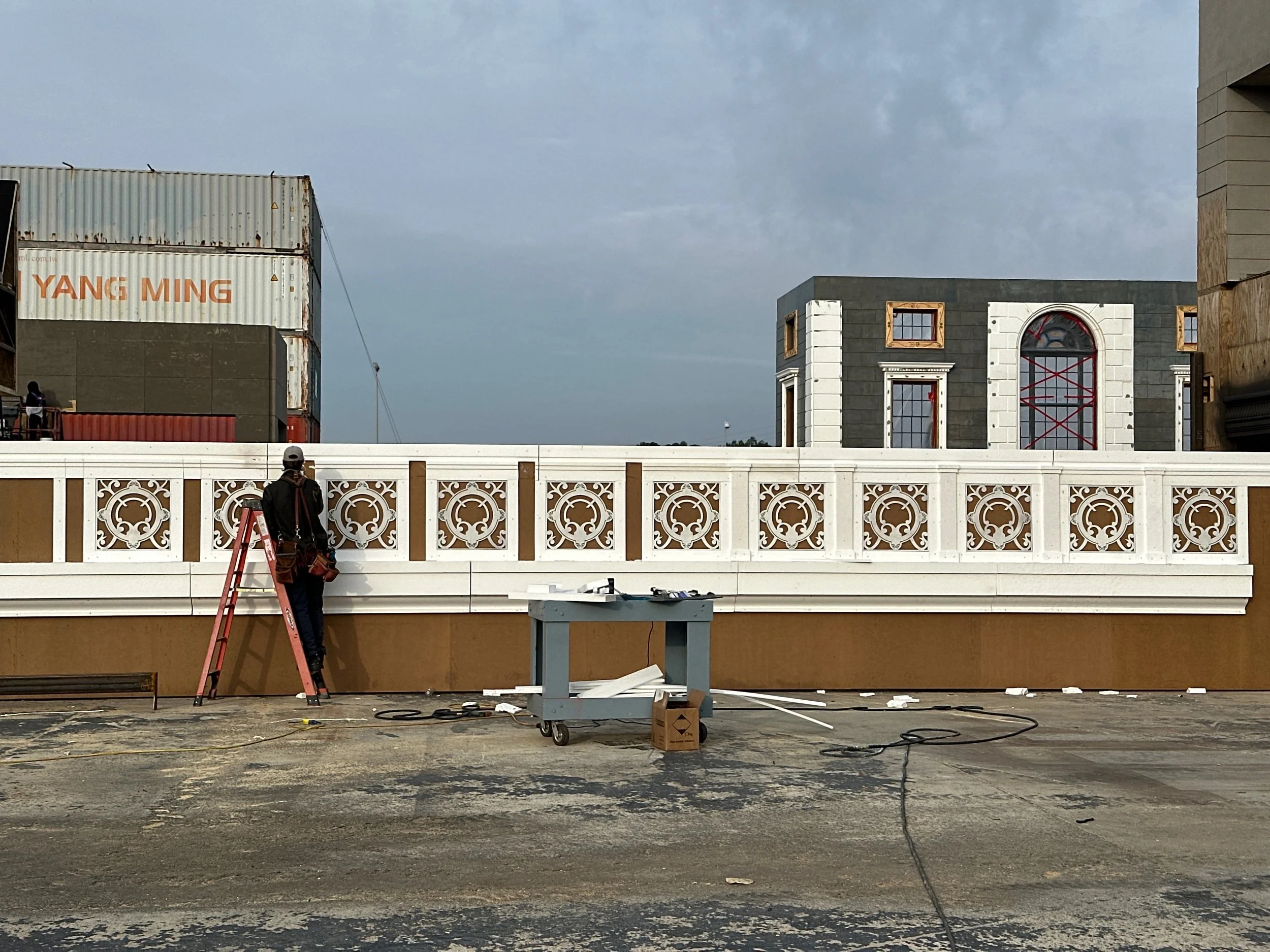 Construction worker working on a white decorative railing on a rooftop, with buildings under construction and cloudy sky in the background.