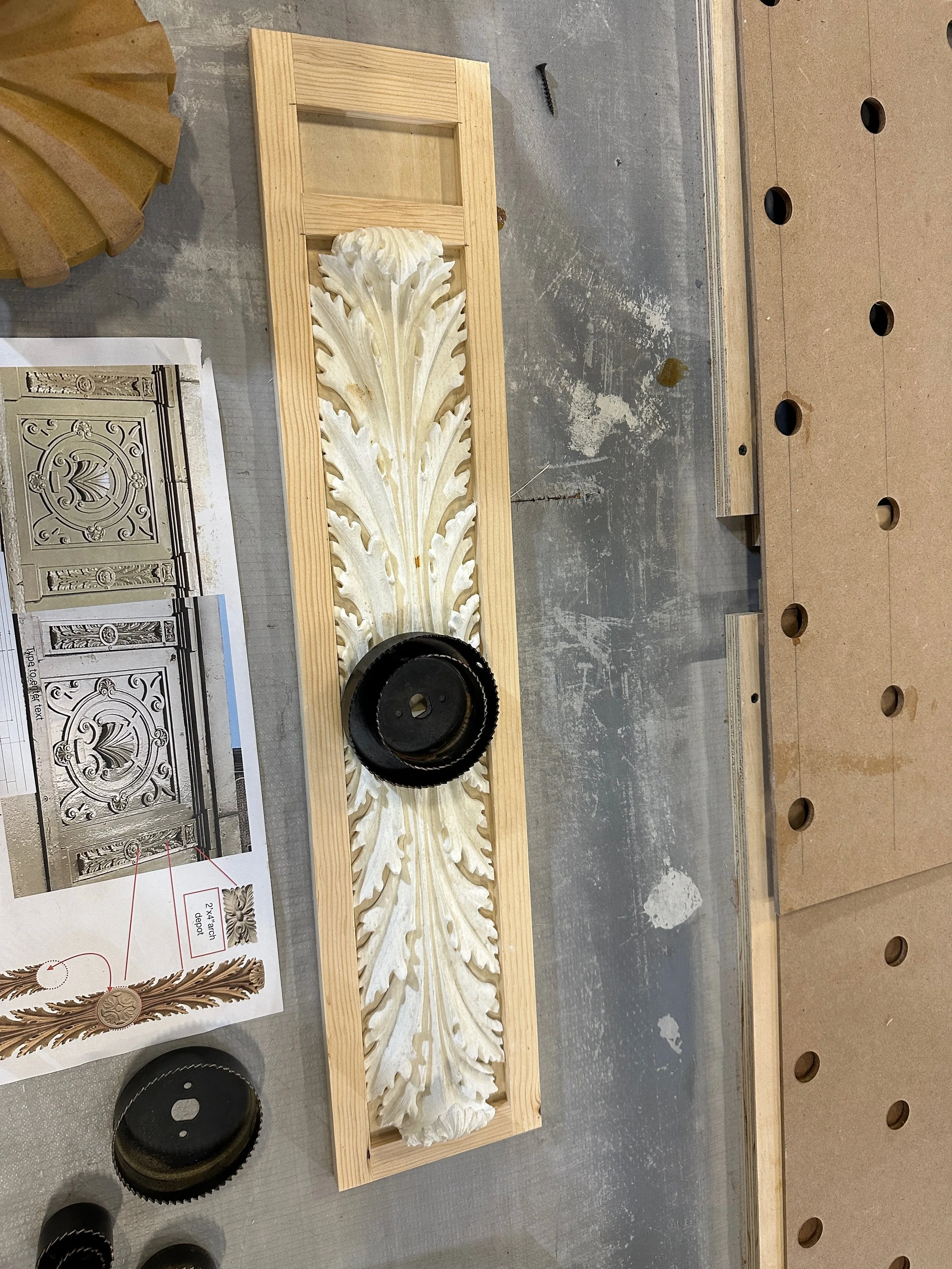 A woodworking project featuring a rectangular wooden frame with an ornate carved design resembling leaves, and black circular saw blades on a workbench. There are also printed design templates and a section of pegboard with holes.