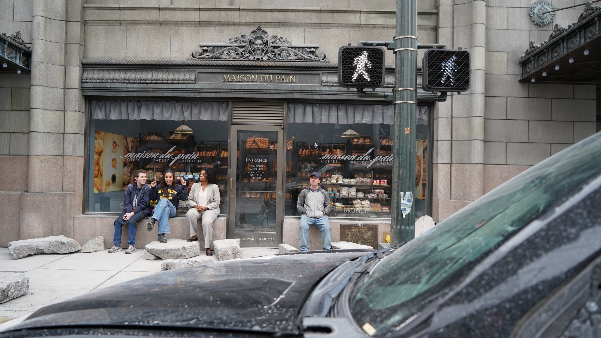 Group of four people sitting on stone blocks outside a bakery called Maison du Pain, with a street and cars in the foreground and traffic lights showing walk signals.