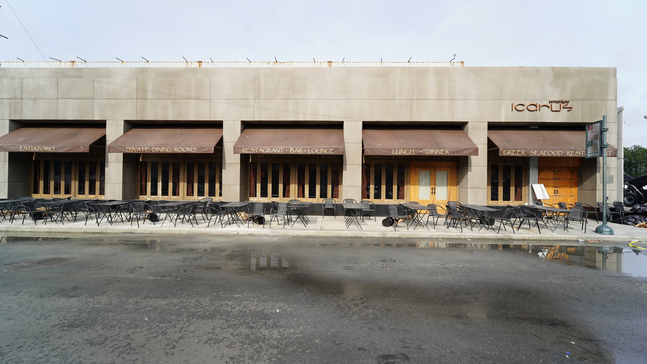 Exterior view of a restaurant with outdoor seating, black tables and chairs, and awnings labeled "Estatorio," "Private Dining Rooms," "Restaurant Bar Lounge," "Lunch Dinner," and "Greek Seafood Rest" on a sunny day with reflections in puddles on the 