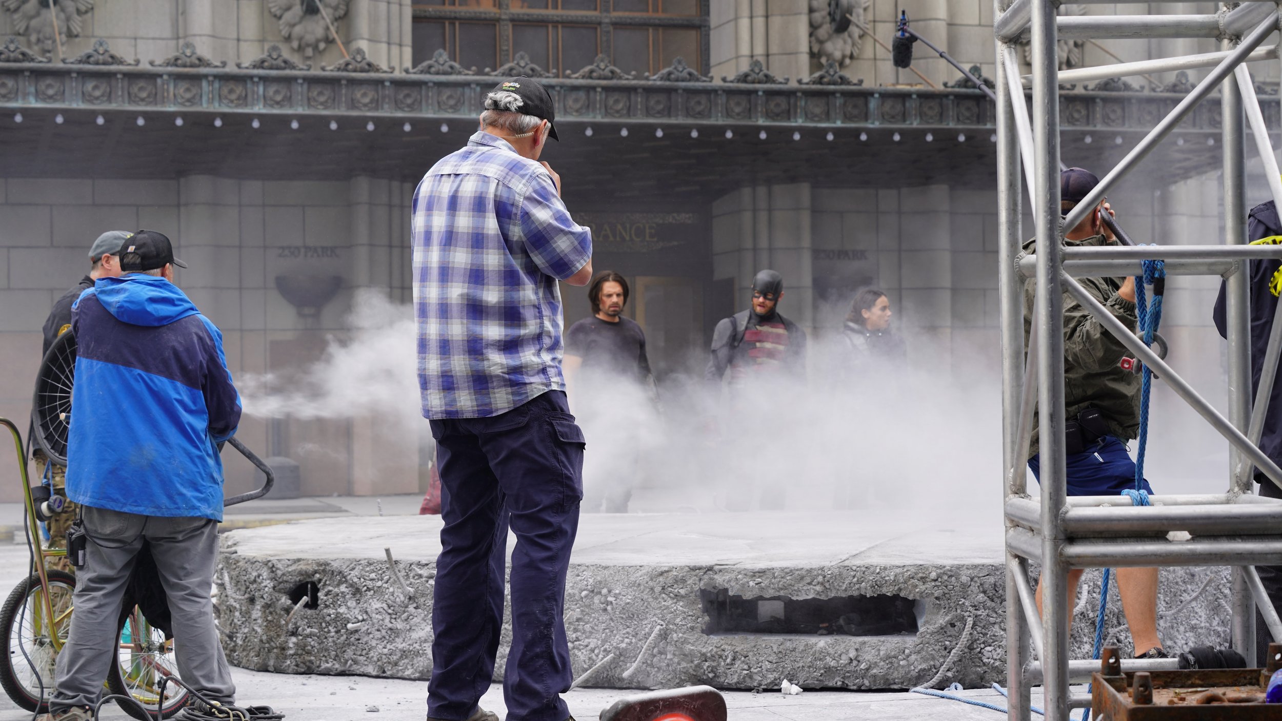 Construction workers working on a city sidewalk with dust and scaffolding, some pedestrians observing in the background.