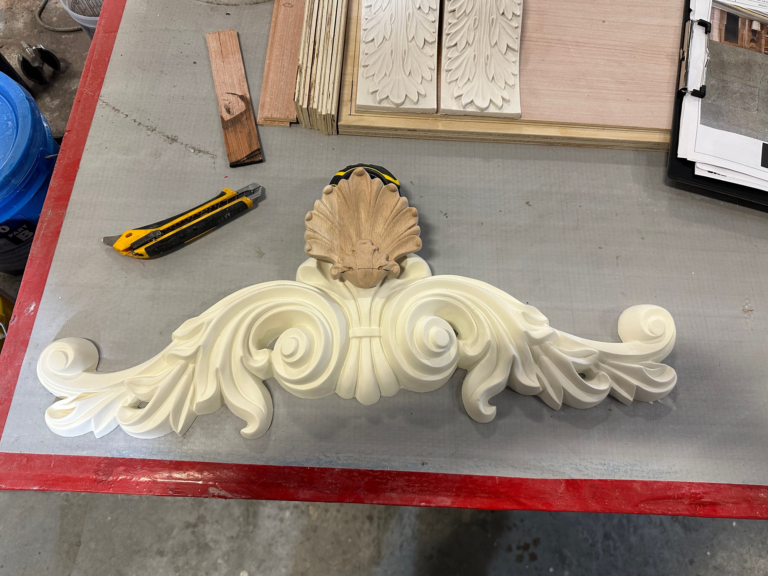 Wooden ornamental carving with scroll and leaf designs on a work table surrounded by woodworking tools and materials.