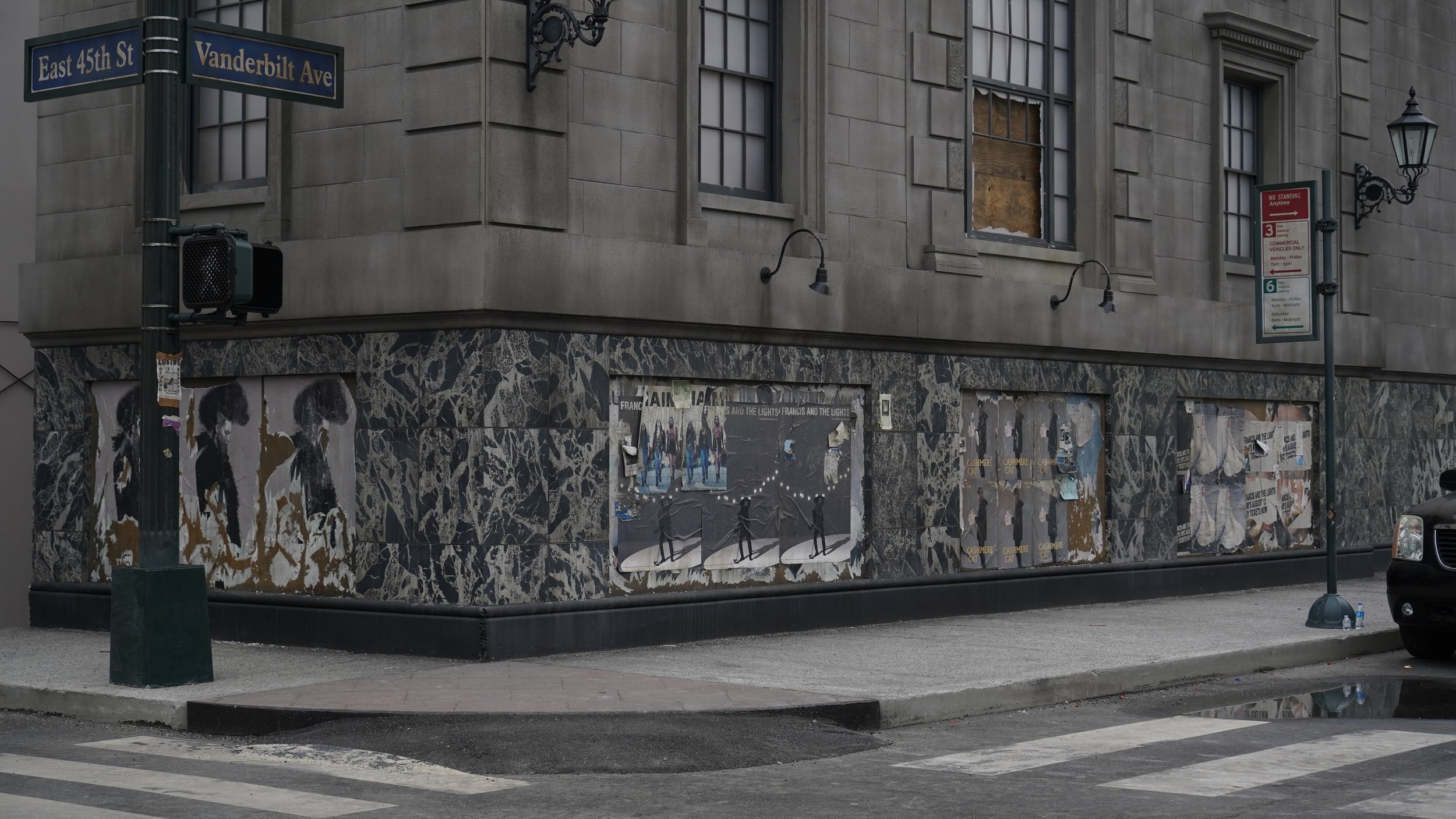 Street corner at East 45th Street and Vanderbilt Avenue with a building featuring boarded-up windows and street posters, a traffic light, and street signs.