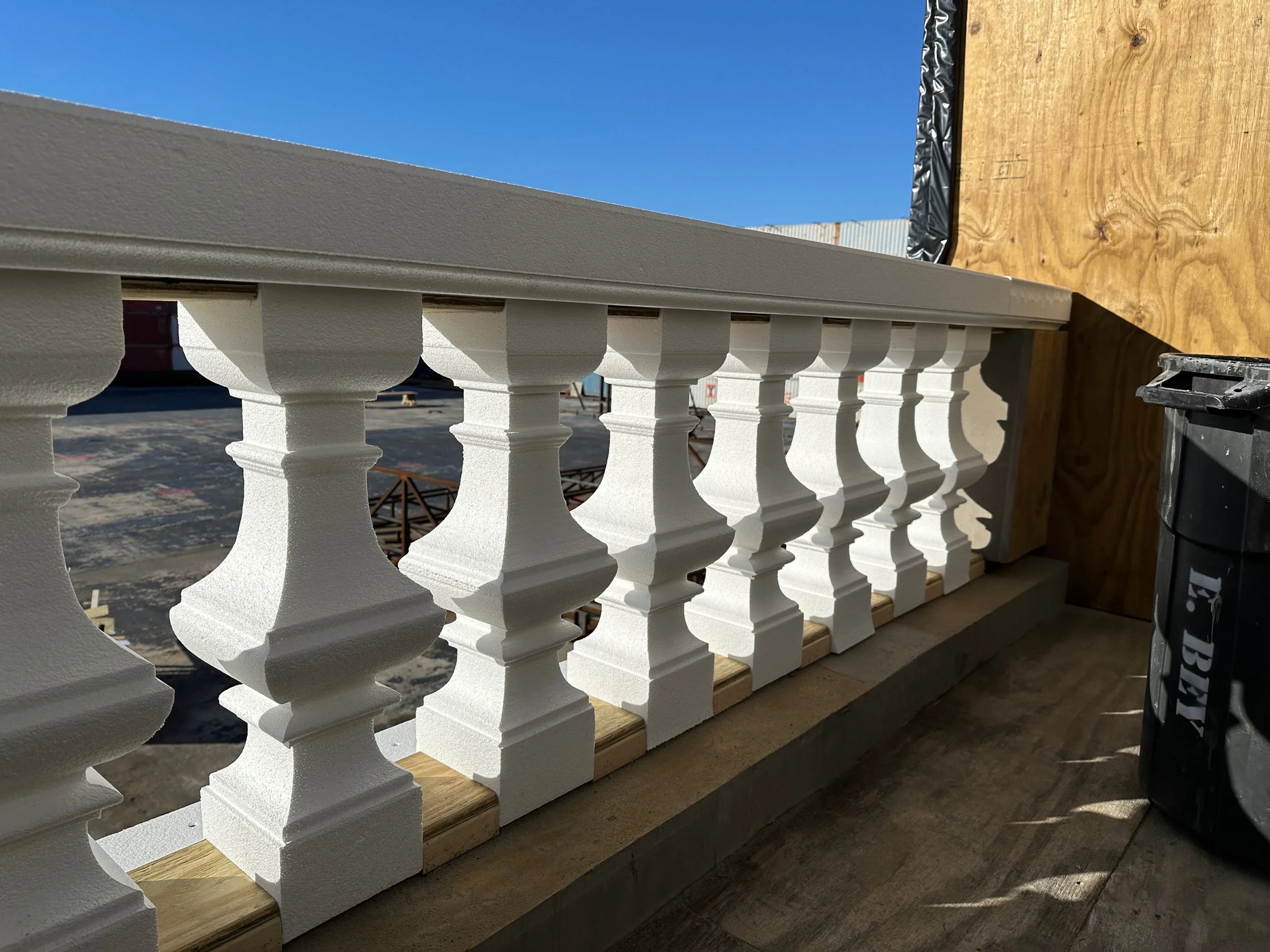 Close-up of a white decorative railing with balusters, mounted on a wooden balcony with a blue sky background.