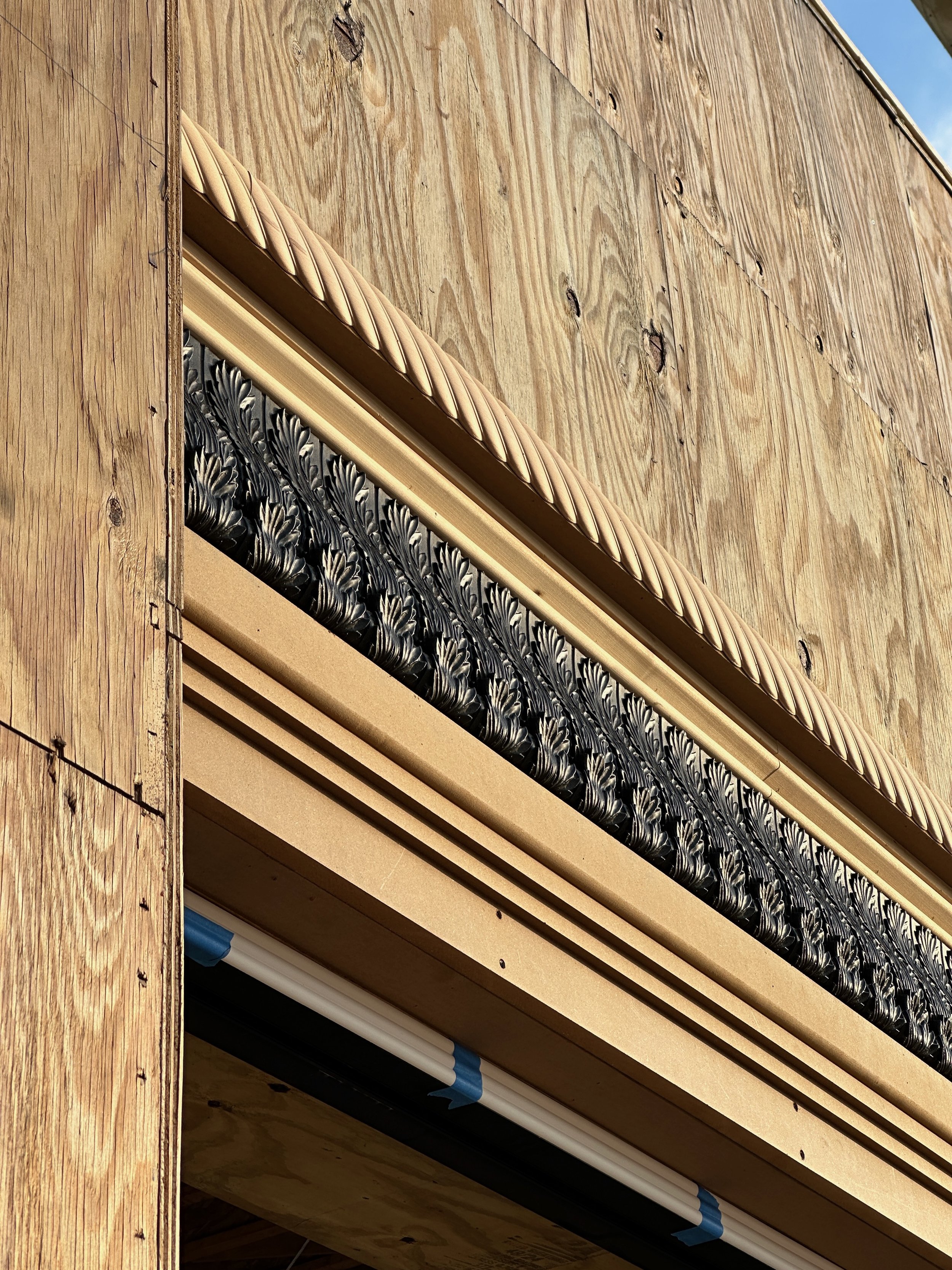 Close-up of a building under construction, showing a wooden exterior wall with decorative molding, black patterned trim, and insulation material.