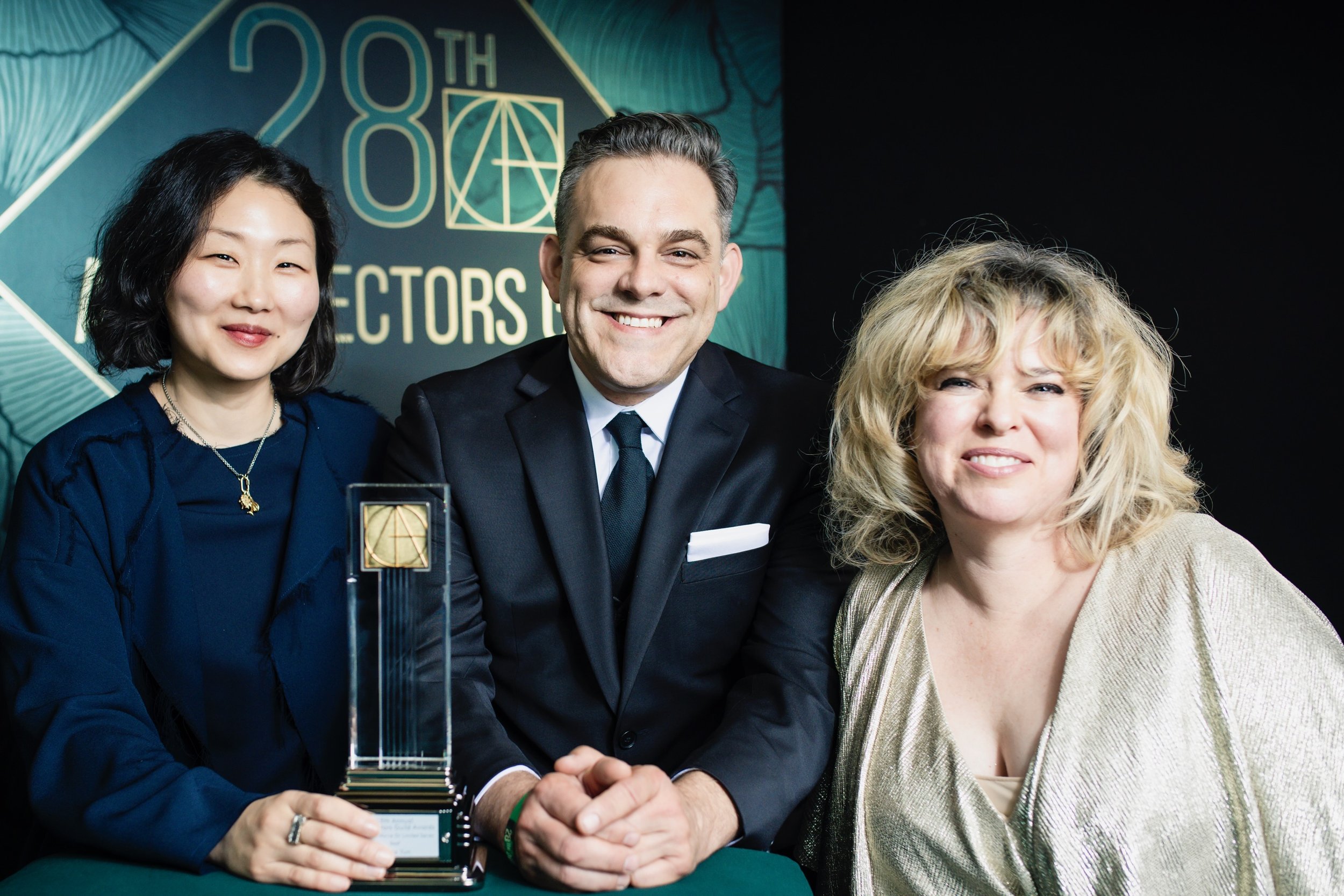 Three people smiling at an award ceremony, one woman holding a trophy, with a sign in the background that reads '28th Directors'.