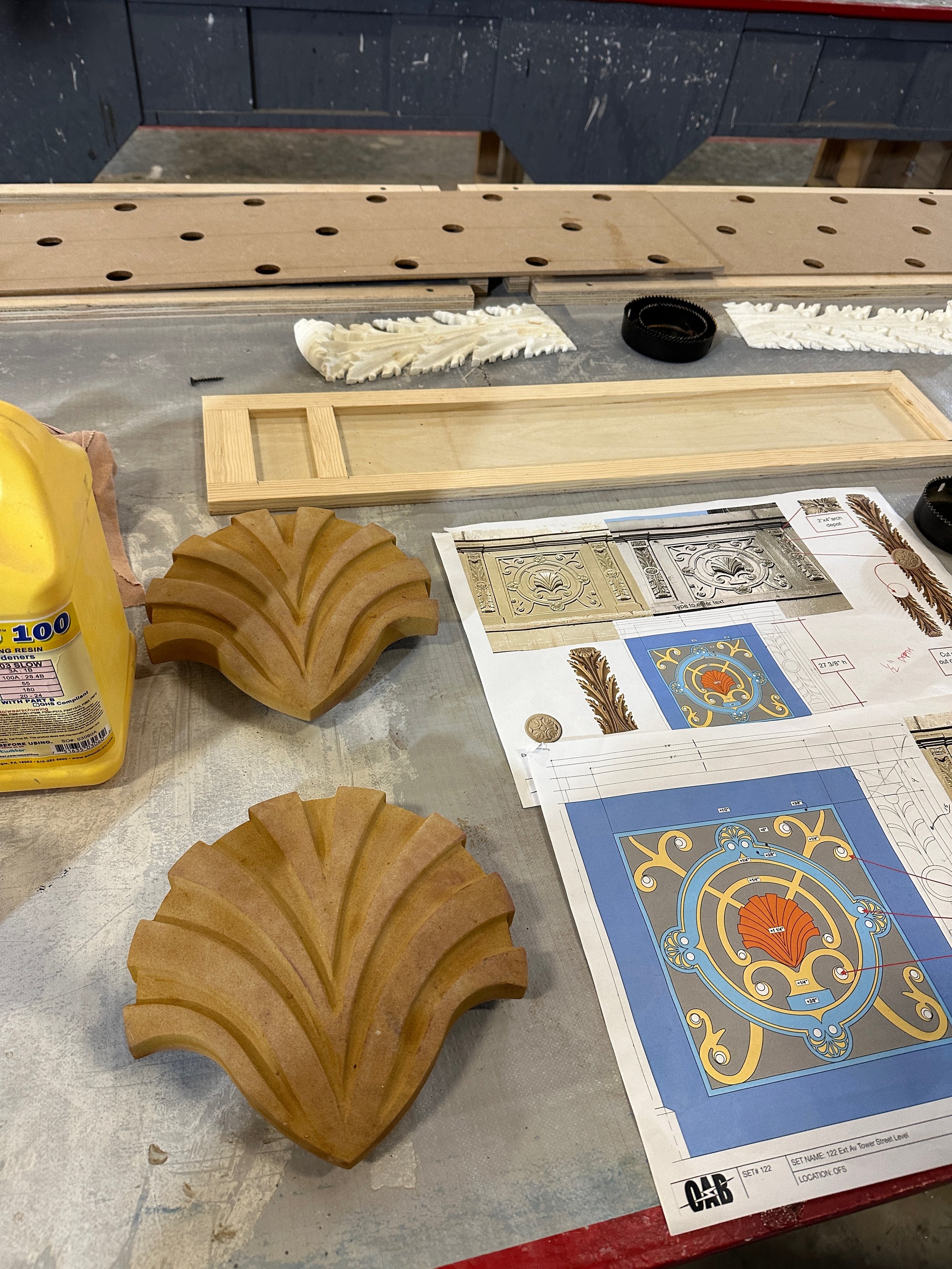 Workshop table with architectural details, blueprints, carved wooden embellishments, and tools.