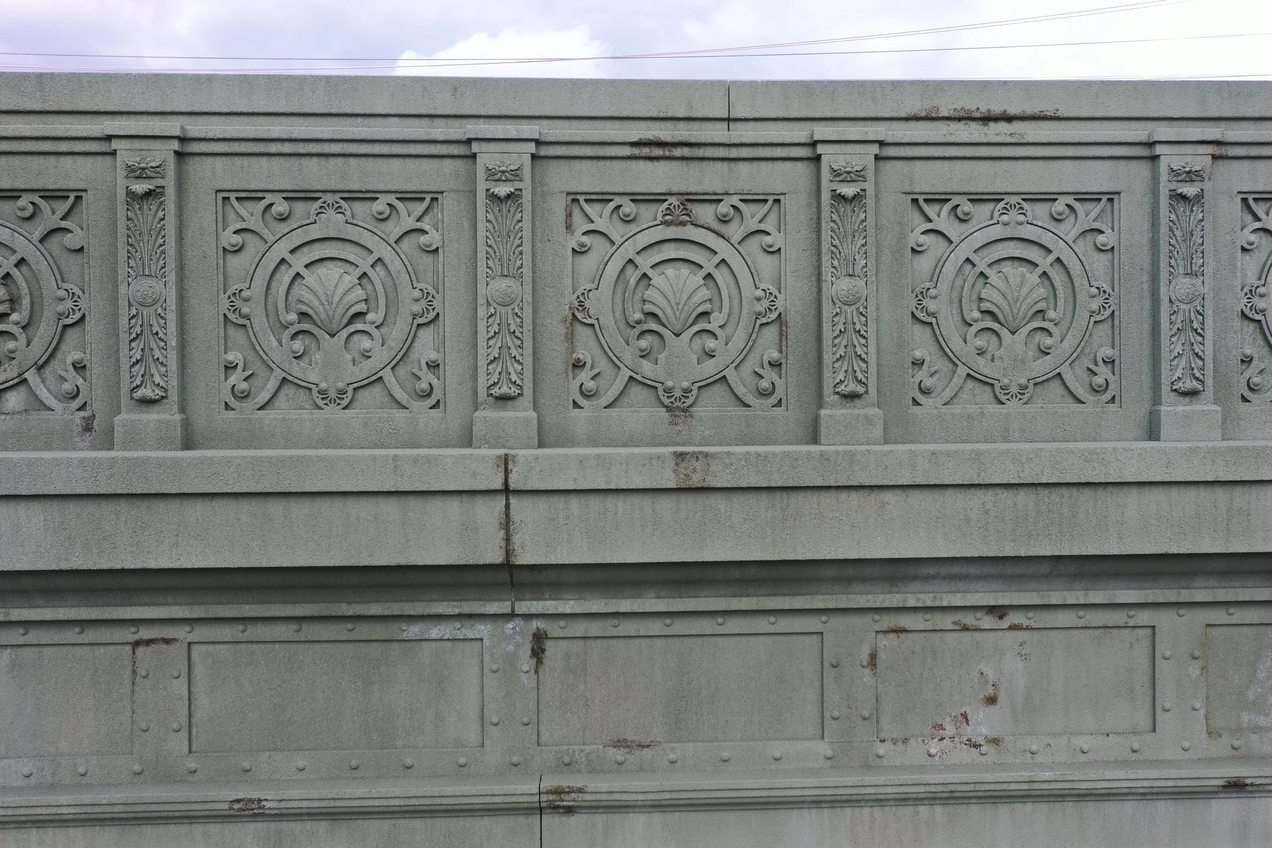 Gray concrete bridge railing with intricate decorative patterns, including floral motifs and circular designs.