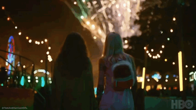 Two girls walking at night in a decorated outdoor space with string lights and fireworks in the background.