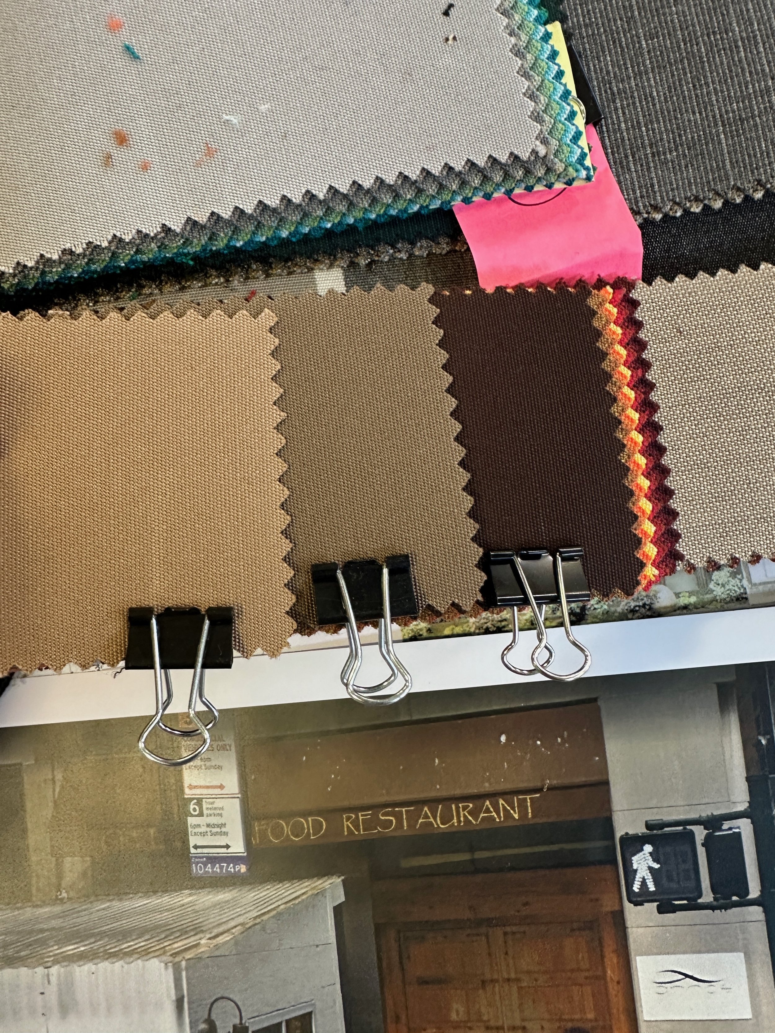 Display of fabric swatches with various colors and patterns, held by black binder clips on a white surface exterior to a restaurant.