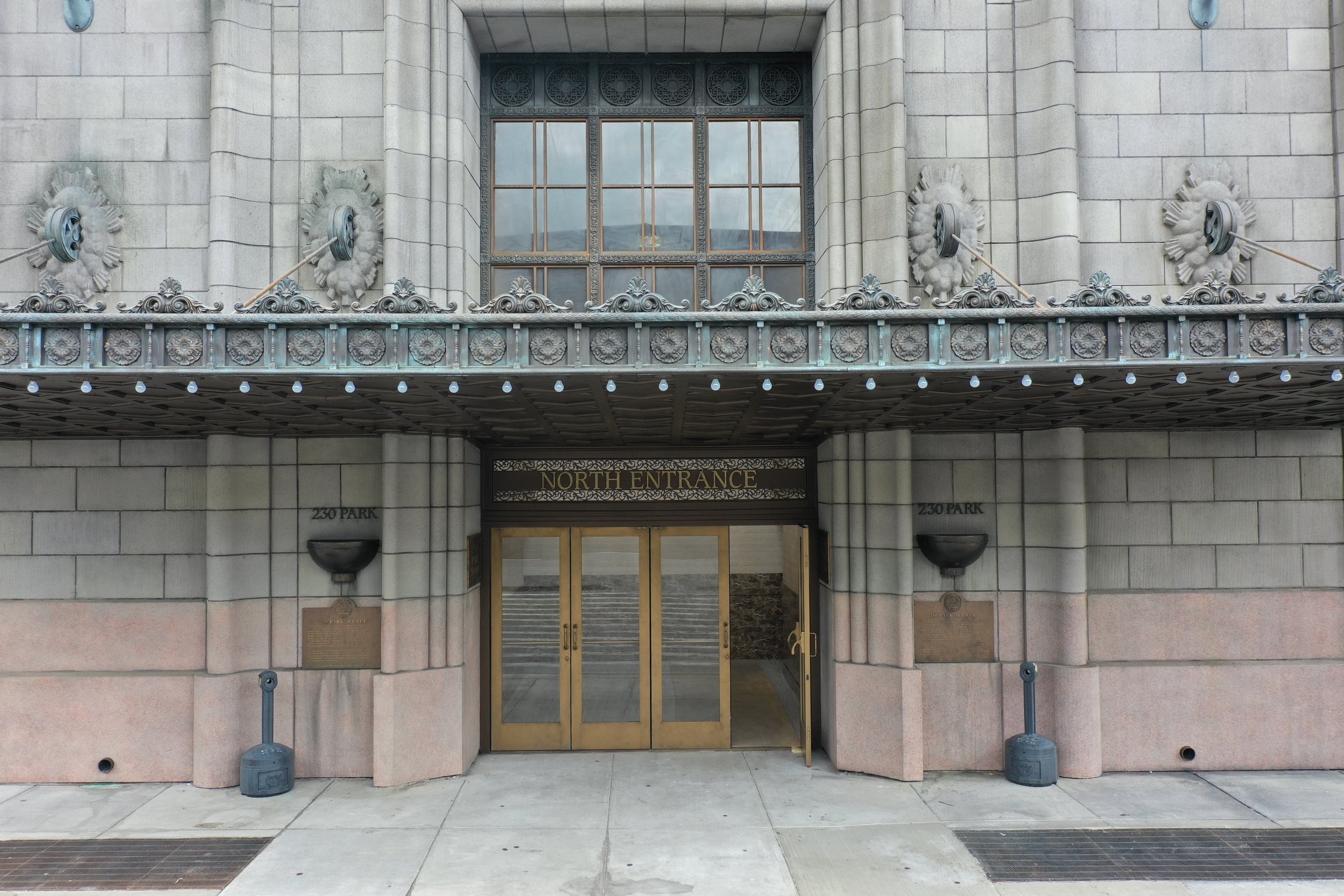 Entrance to a building with a sign that reads 'North Entrance'. The building has a stone facade, decorative metalwork, and large windows. There are two black wall-mounted light fixtures on either side of the entrance, and the doors are glass with gol