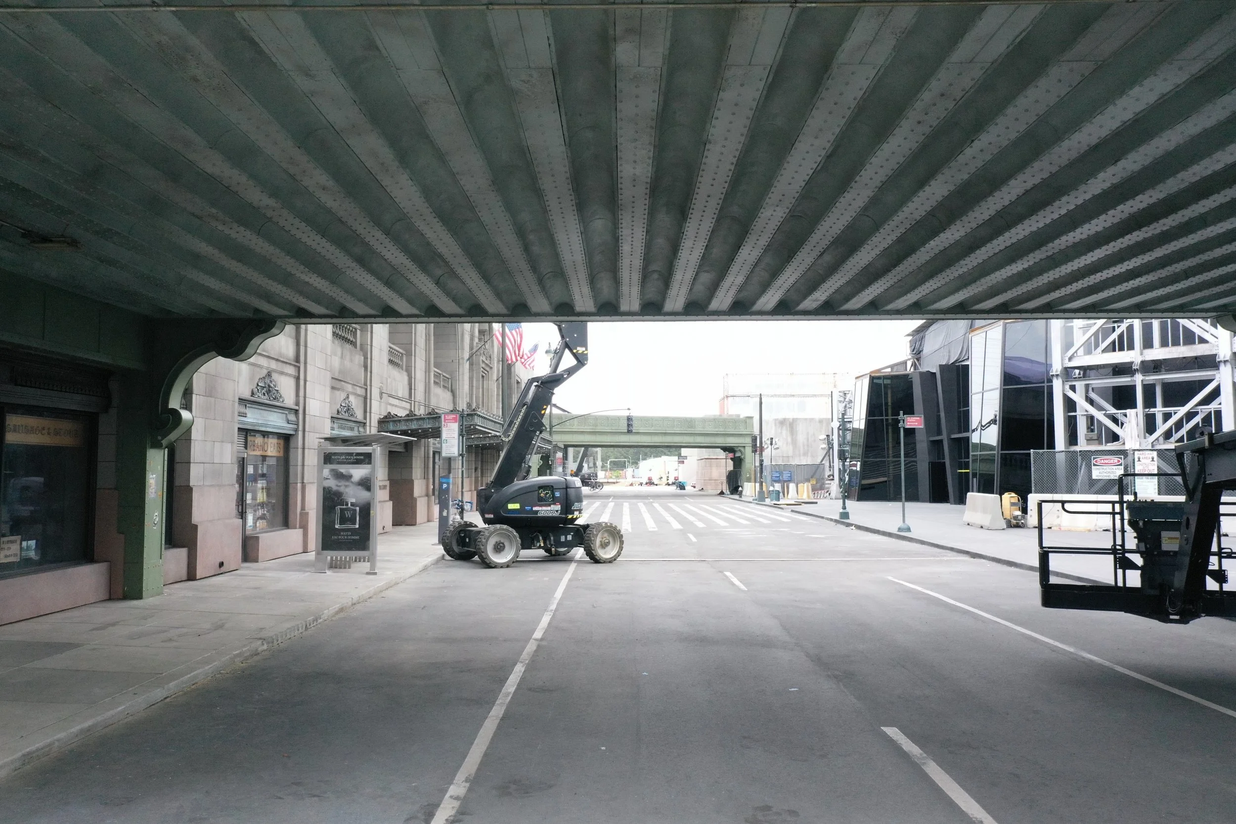 Empty street under a bridge with construction equipment and buildings on both sides, including a black lift and scaffolding.