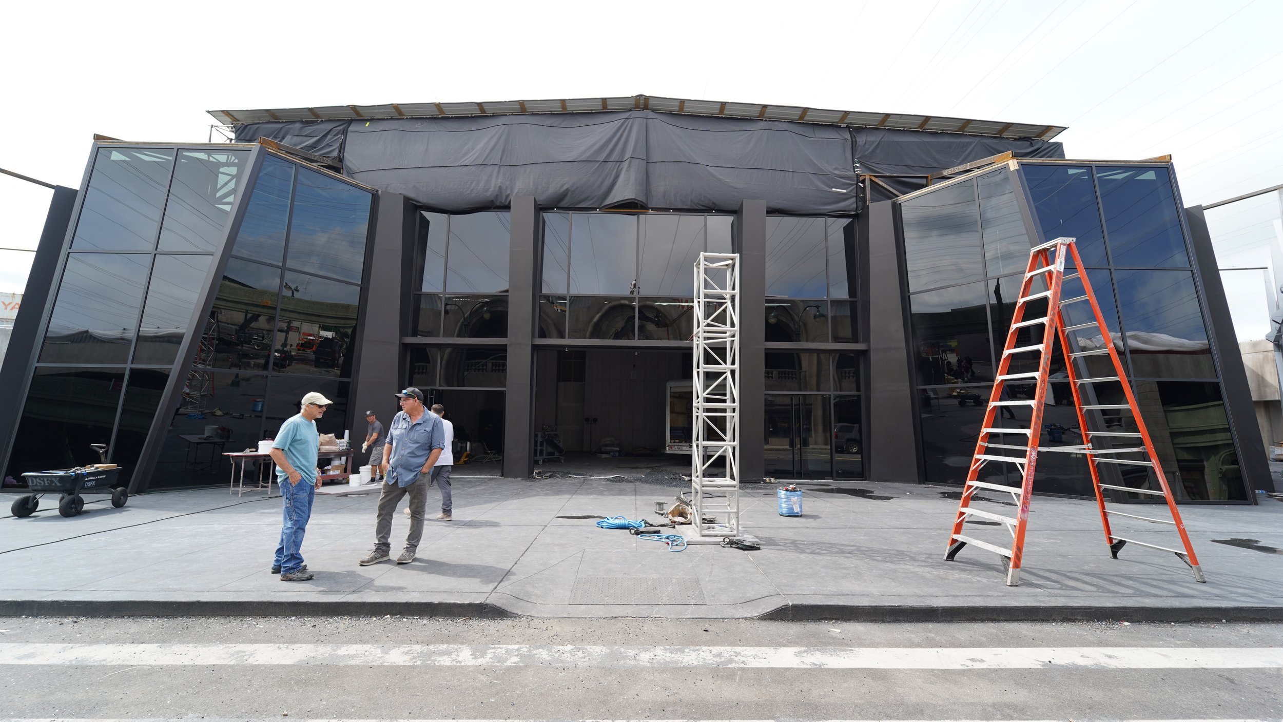 Construction crew working on a modern black building with large reflective glass windows, ladders and scaffolding outside.