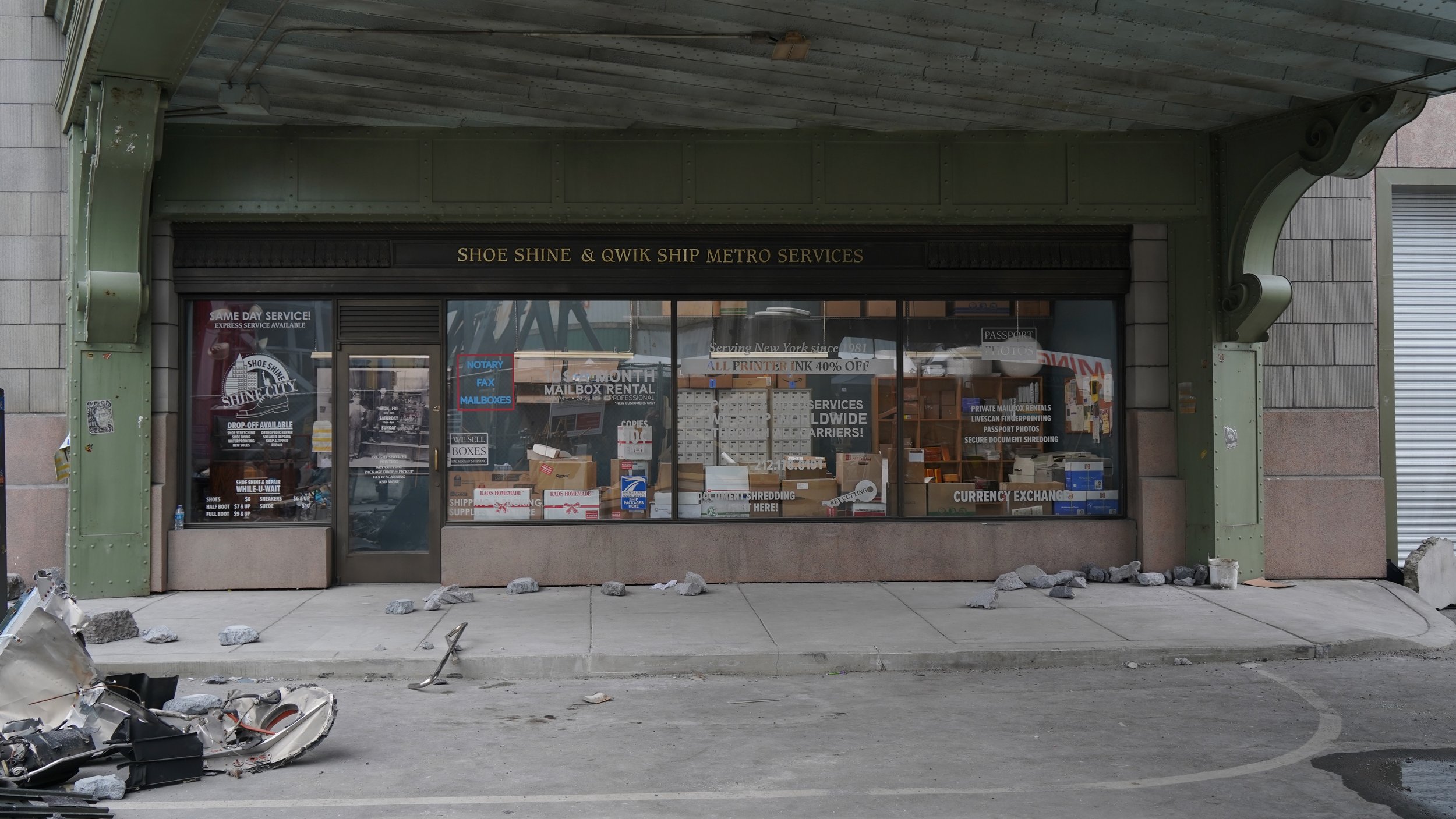 Storefront called "Shoe Shine & Qwik Ship Metro Services" with display window filled with boxes, signs, and advertising for mailbox rentals, document shredding, and printing services. Debris and damaged scooter parts are in front of the store.