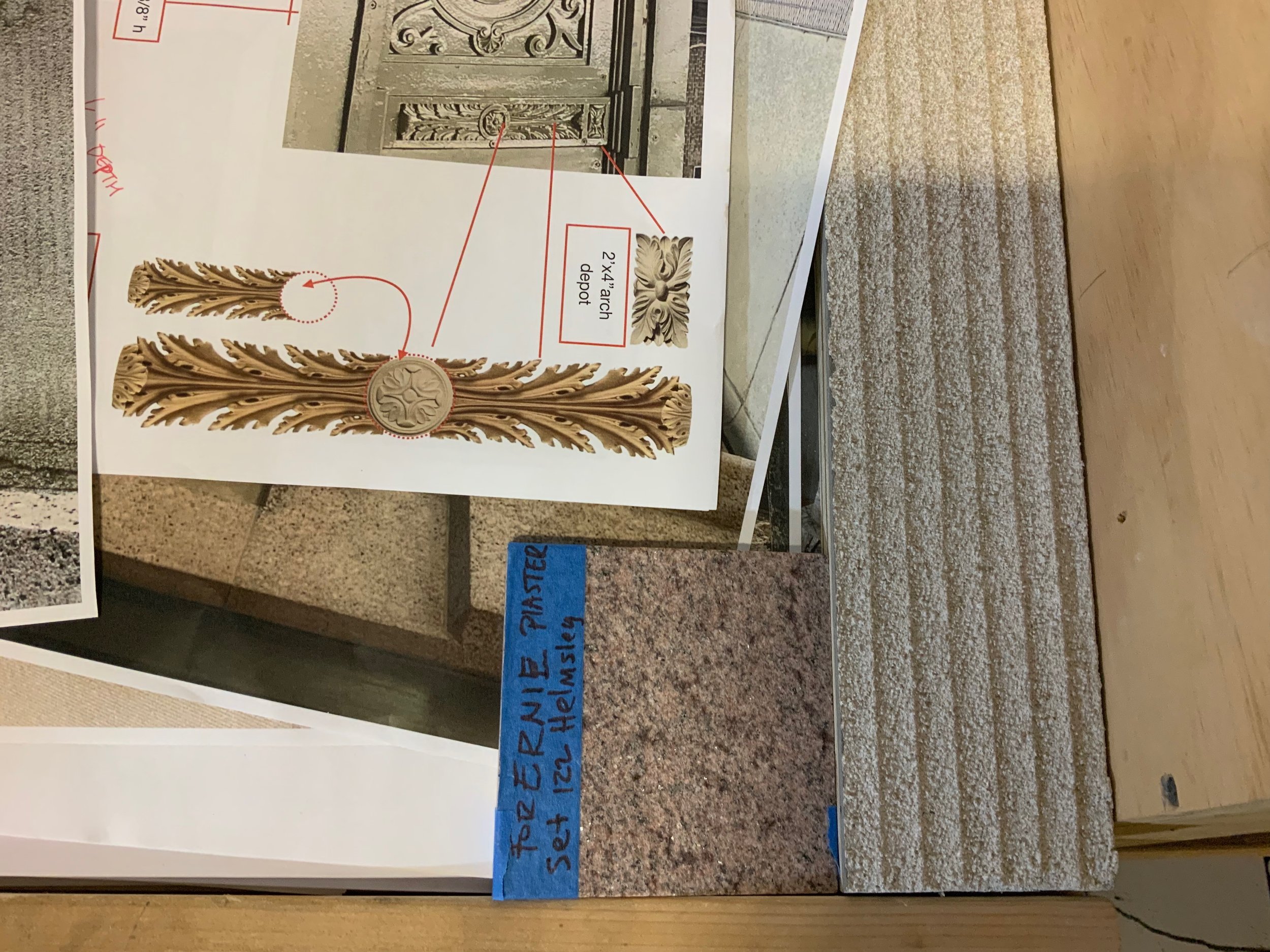 A close-up of a decorative wood carving design for architectural molding, positioned on a table next to a pamphlet with text, and a blue sticky note with handwritten text in blue ink.